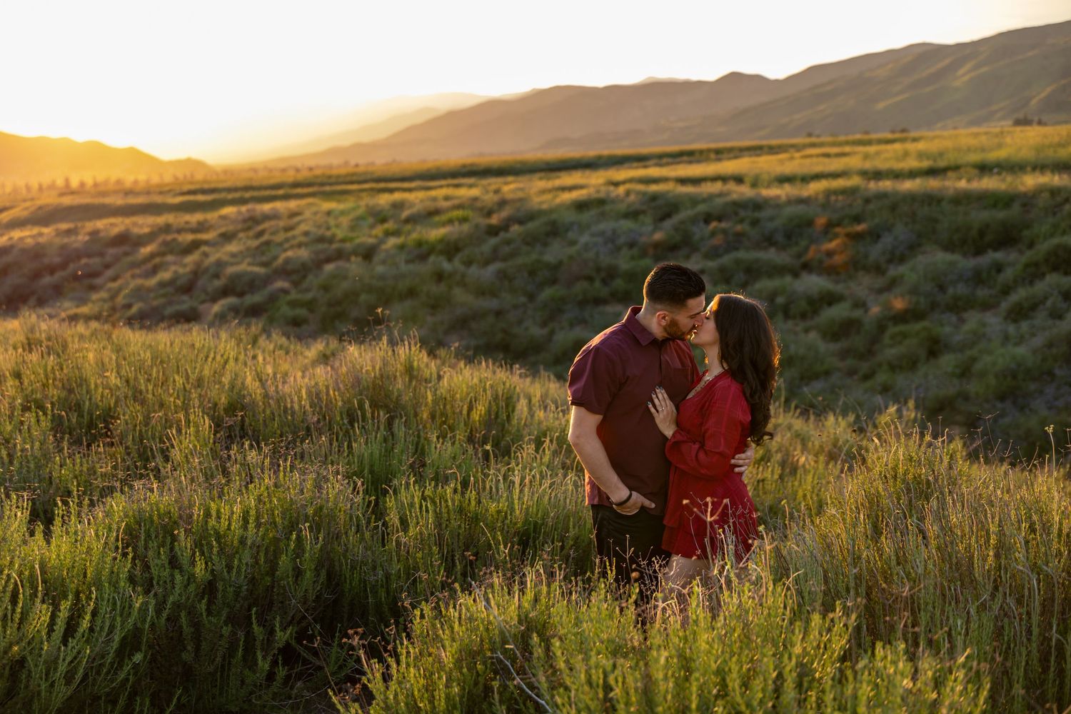 A couple share a romantic moment at sunset in a field of tall grass with mountains in the background in Oak Glen.