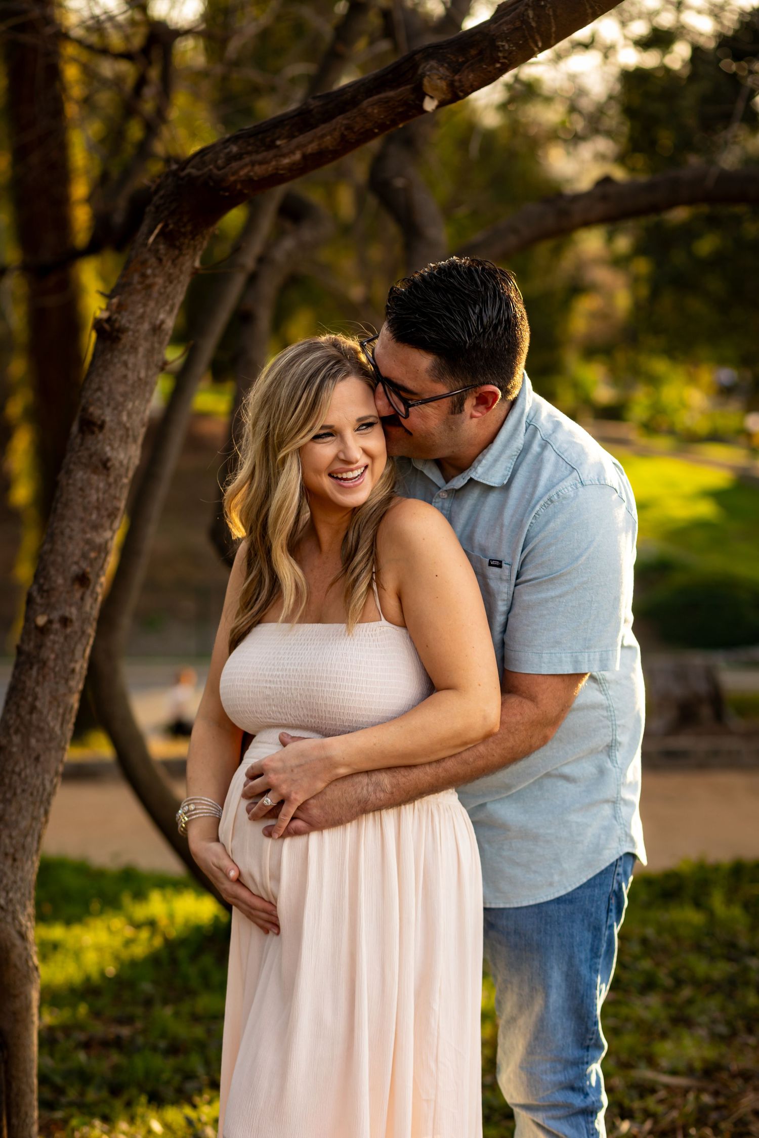 A couple embraces tenderly in a sun-dappled forest setting wearing casual clothing at sunset in Redlands.