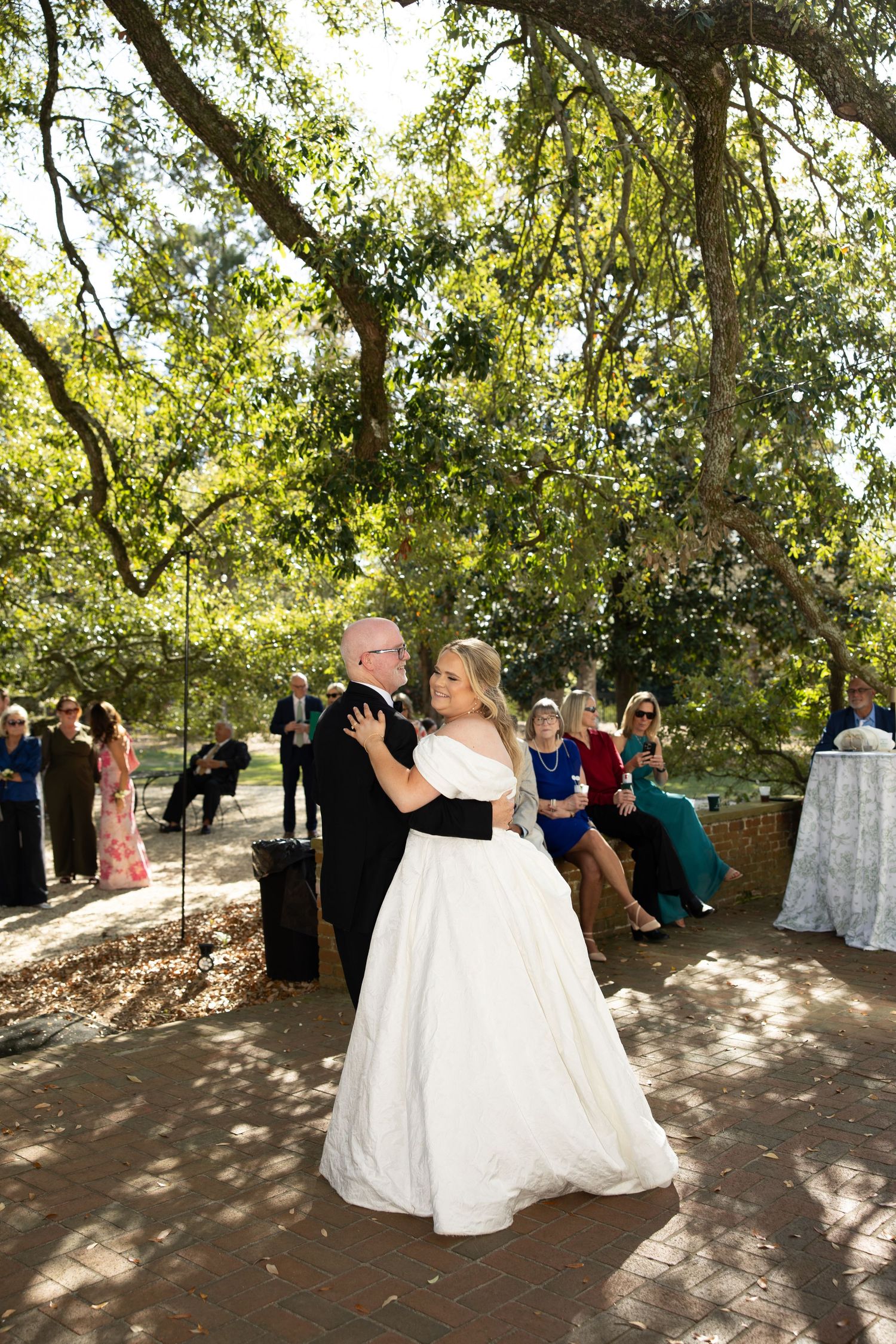 A couple shares a romantic dance under a canopy of trees at an outdoor wedding celebration.