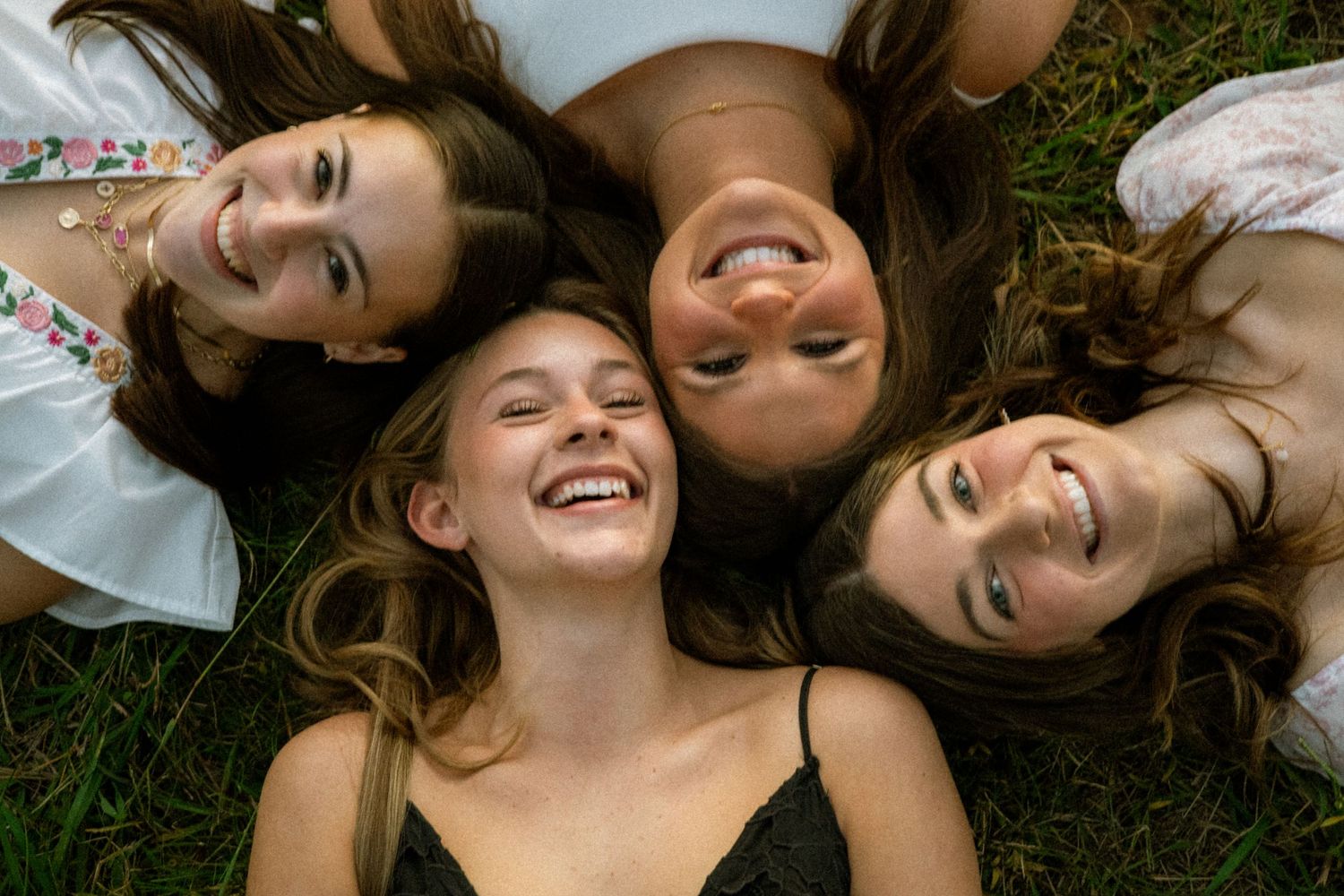 Four friends laying on grass with their heads together forming a circle and laughing.