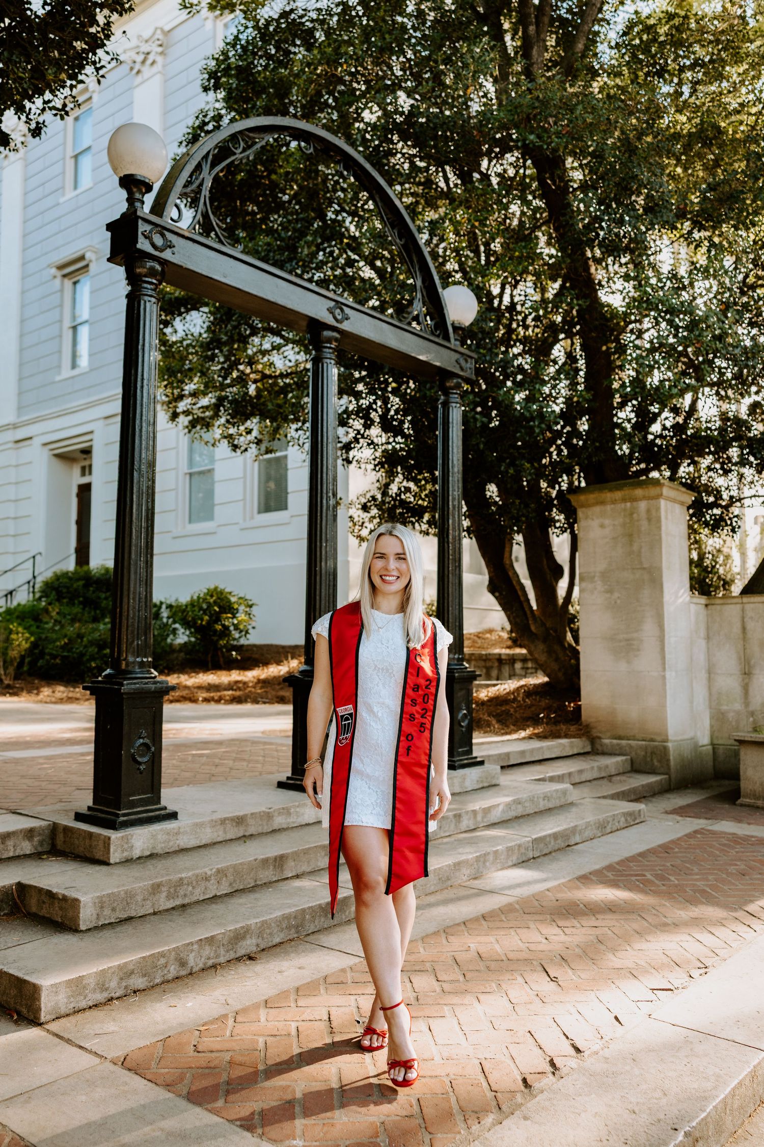Graduate in red stole poses under decorative archway on brick pathway.