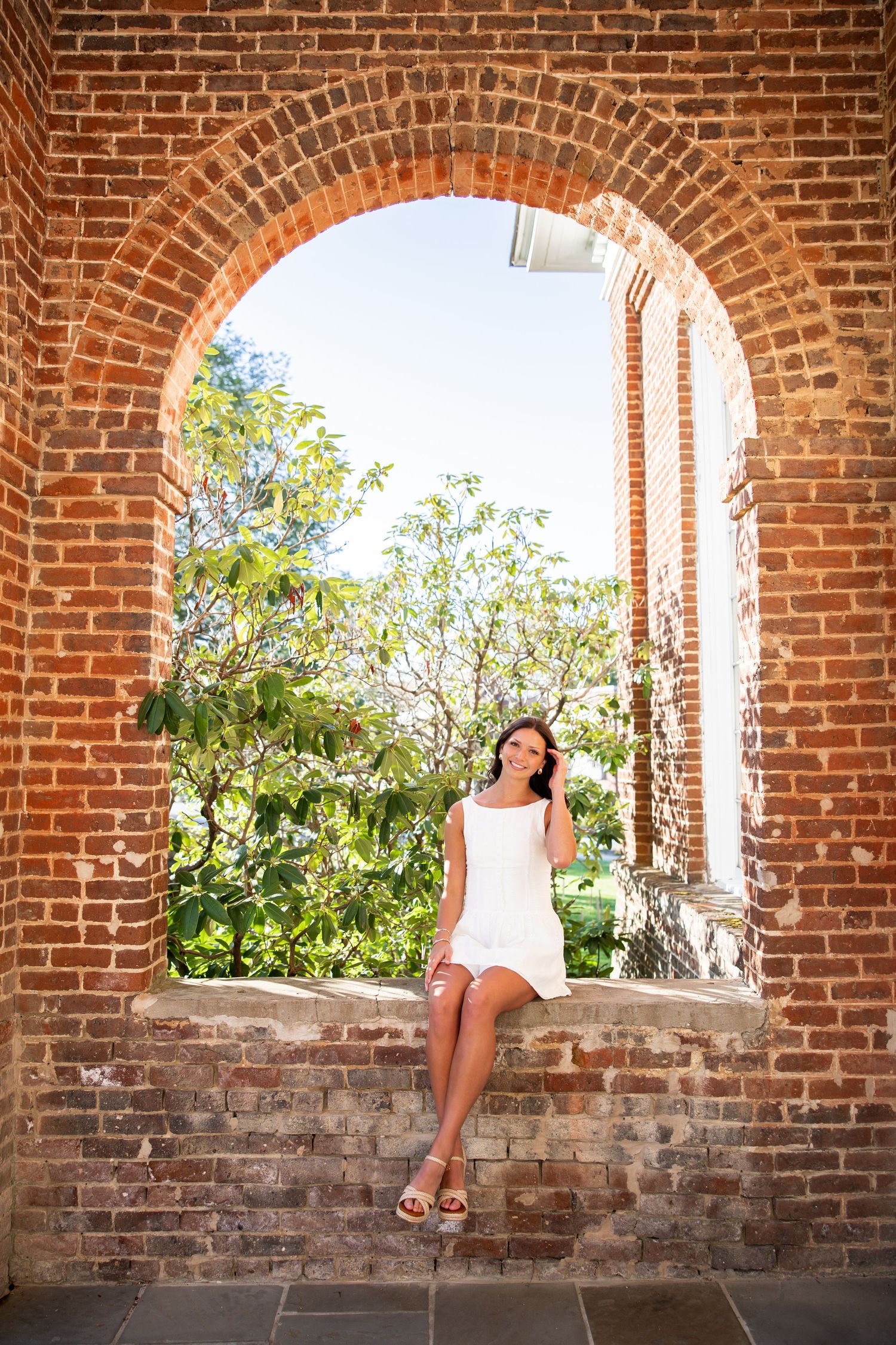 Woman in white dress sitting under brick archway with sunlit garden in background.