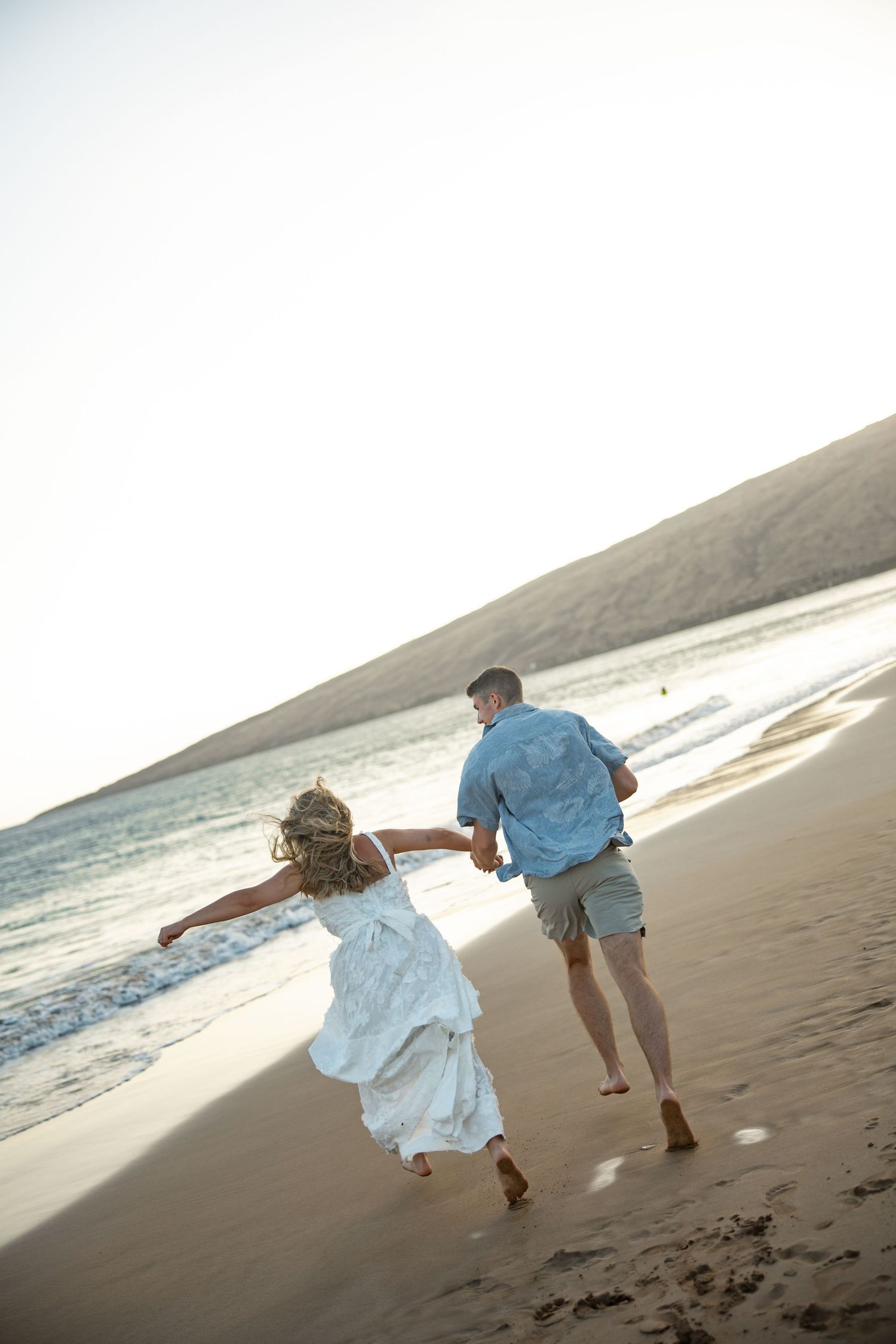 Couple walking and playing together on a sandy beach at sunset with ocean waves in the background.
