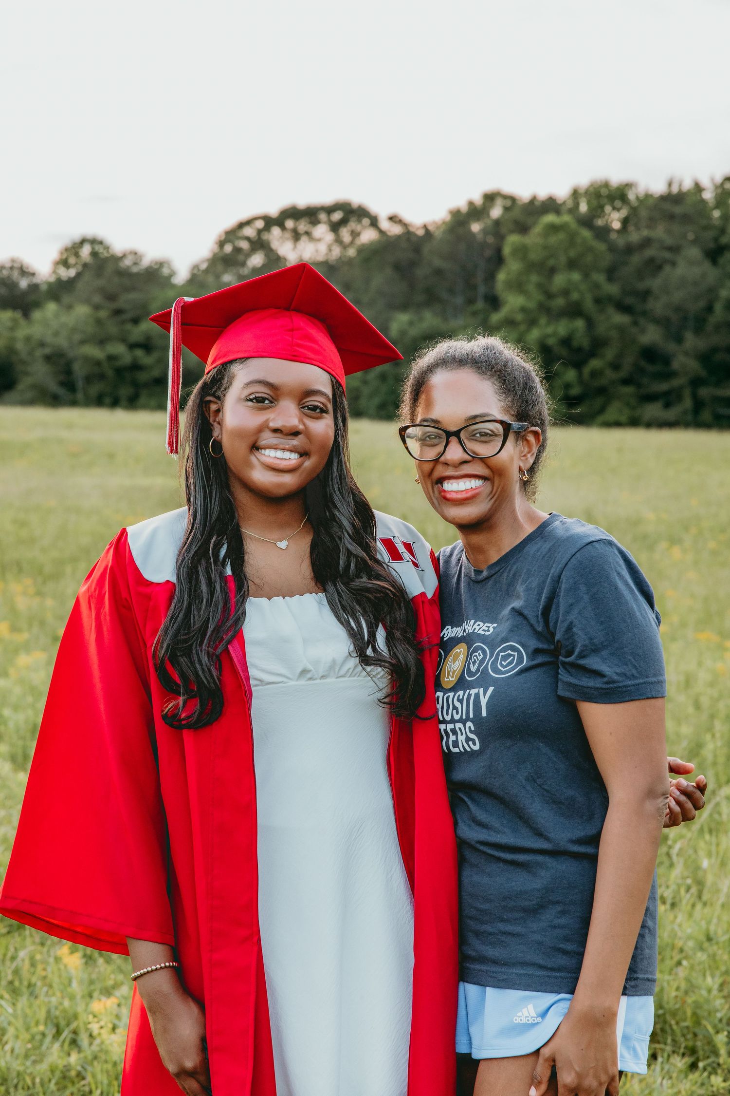 Two people celebrate graduation day together in a grassy field with trees.