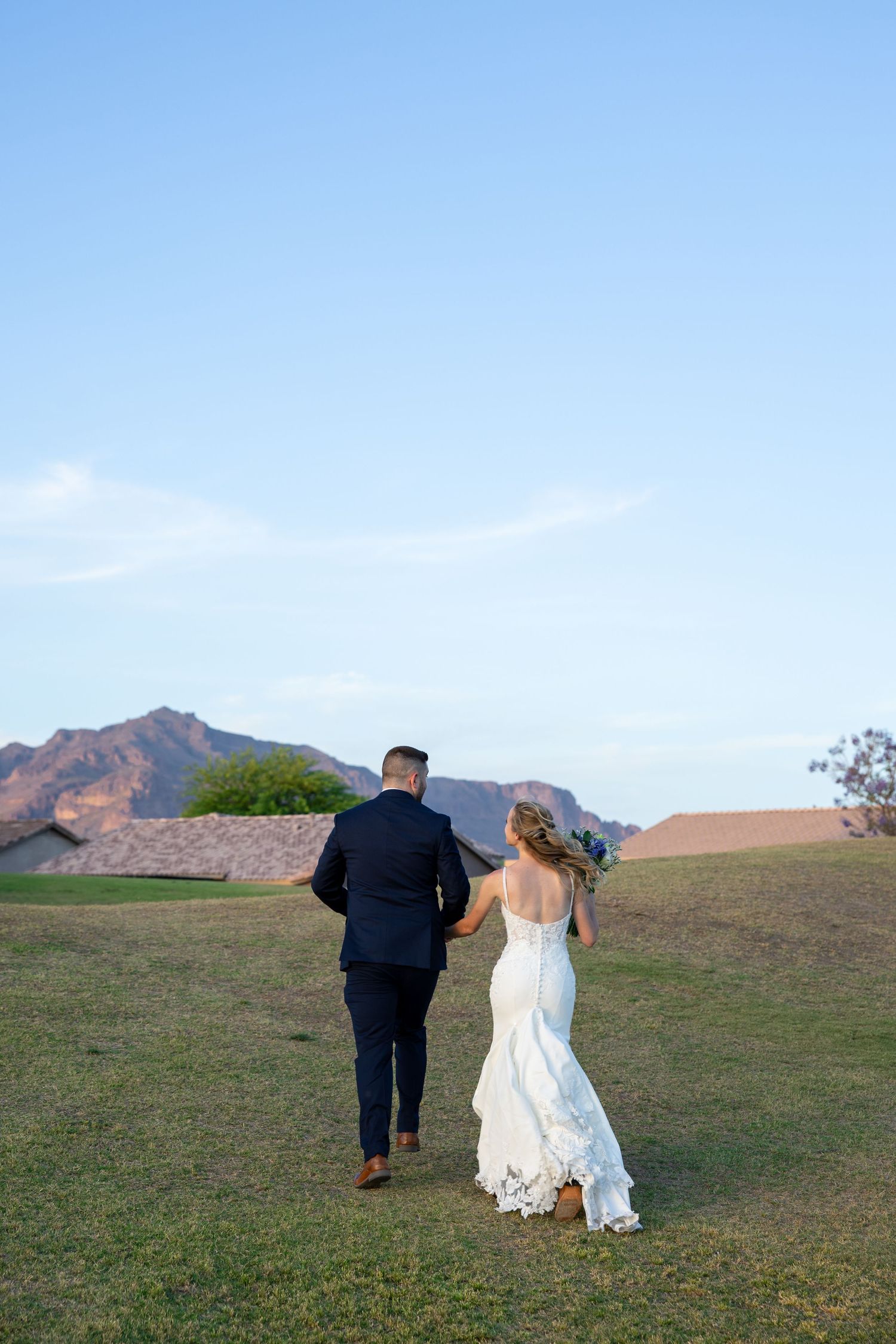 Bride and groom walking hand in hand across green lawn with mountain backdrop at sunset.
