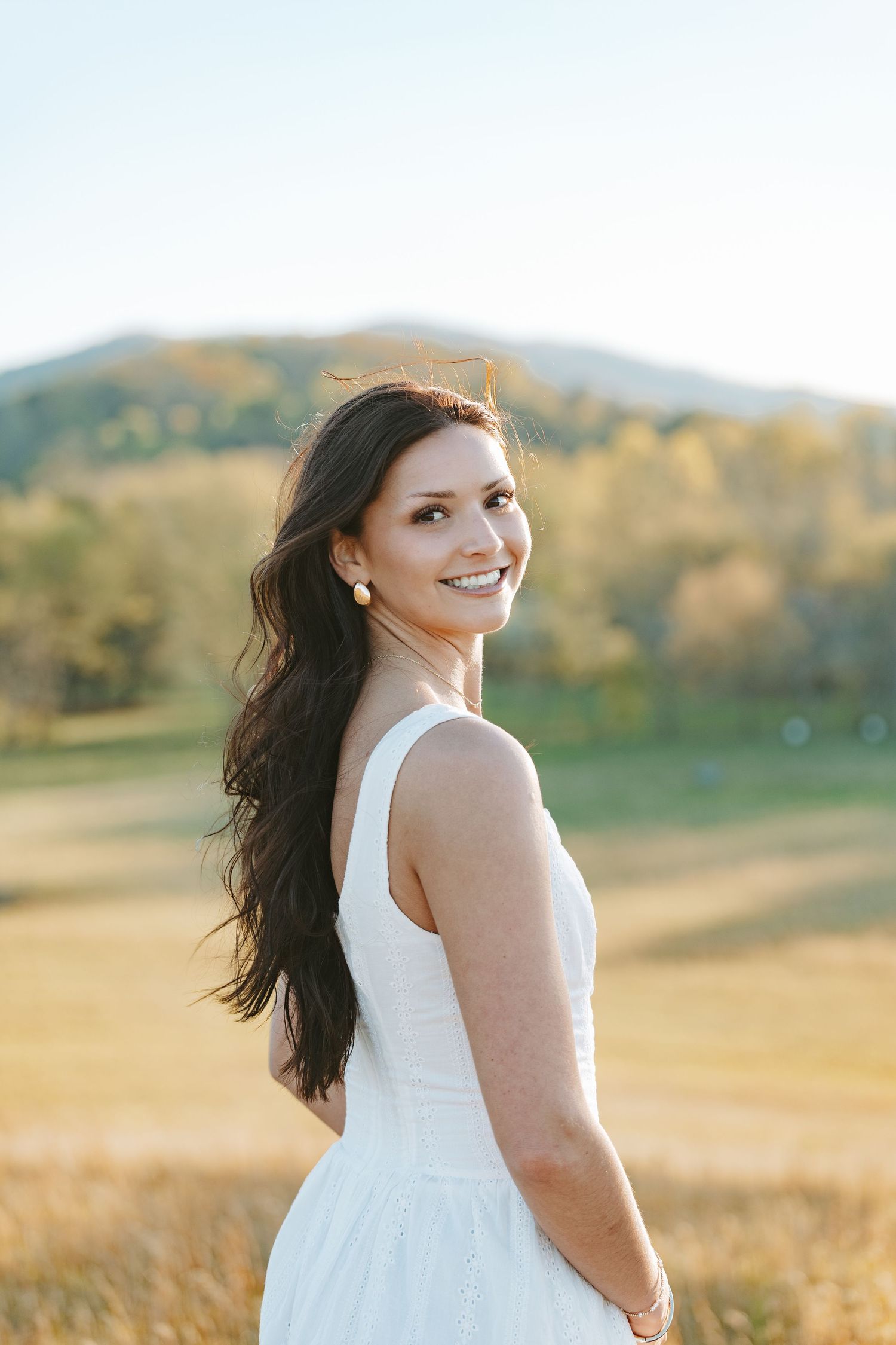 Woman in white sundress poses in golden field with rolling mountains in background.