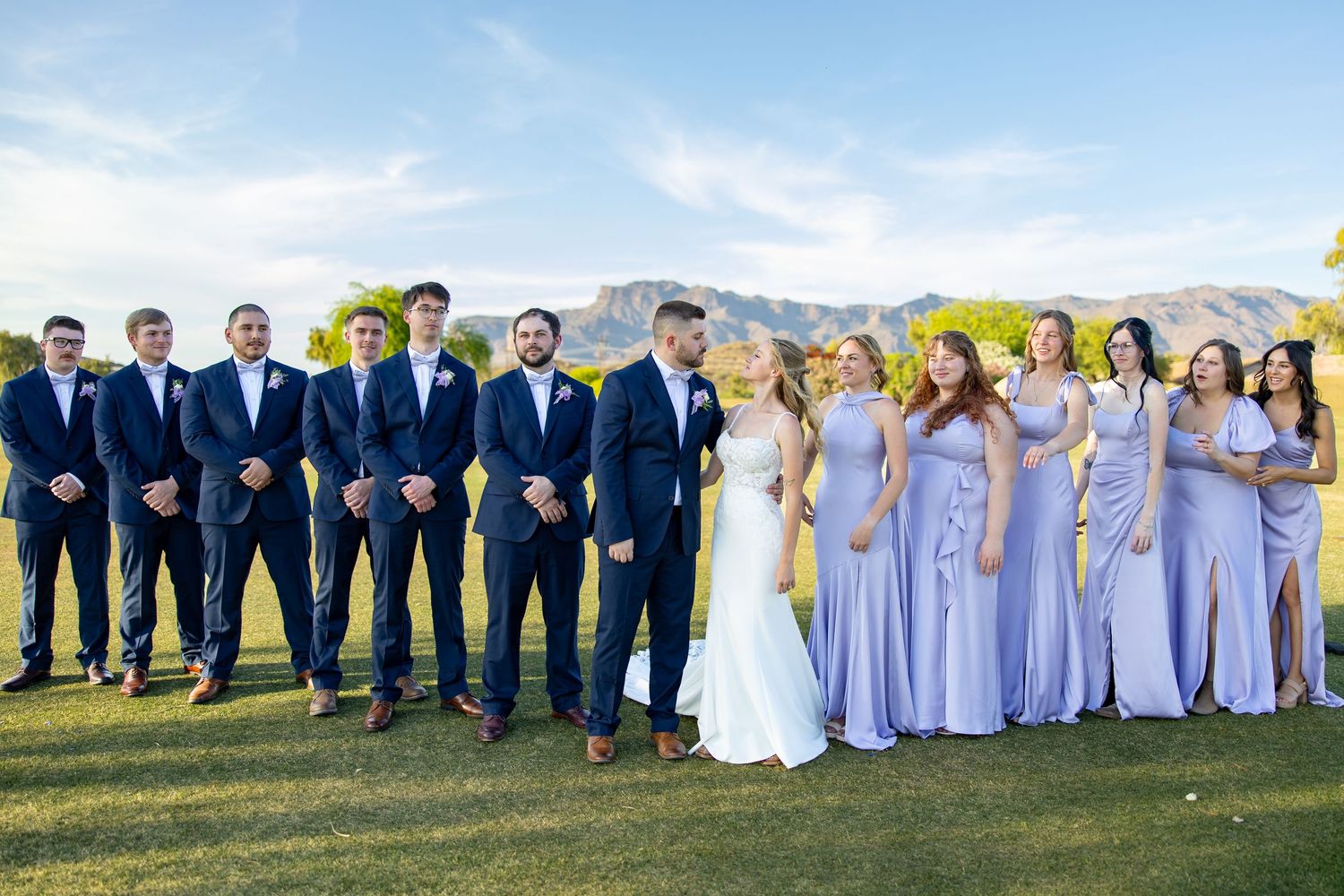 Wedding party poses outdoors with groomsmen in navy suits and bridesmaids in lavender gowns, mountains in background.