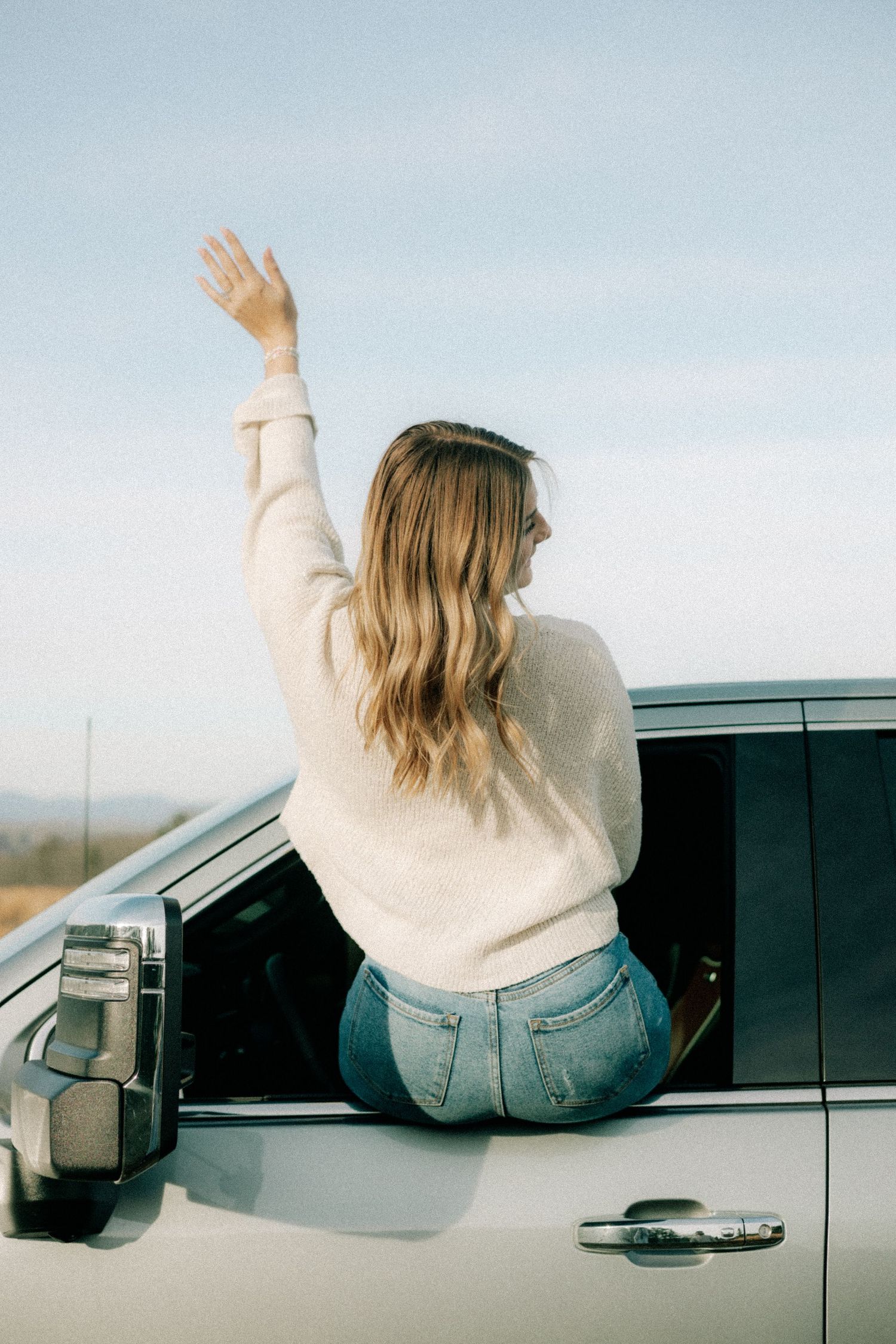 A person in a cream sweater and jeans leans out of a car window with arms raised.