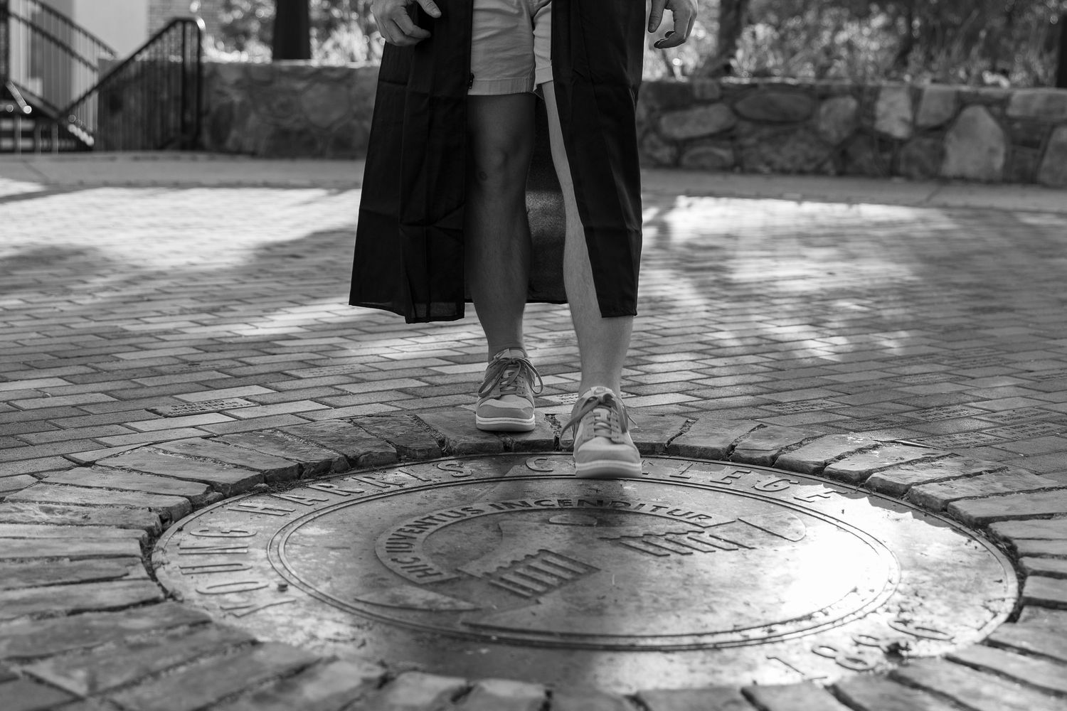 Black and white photo of feet standing on circular university seal embedded in ground.