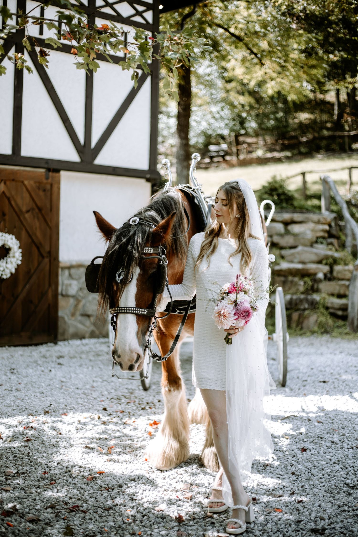 Bride in white dress holding pink flowers stands beside decorated horse at Tudor-style stable.