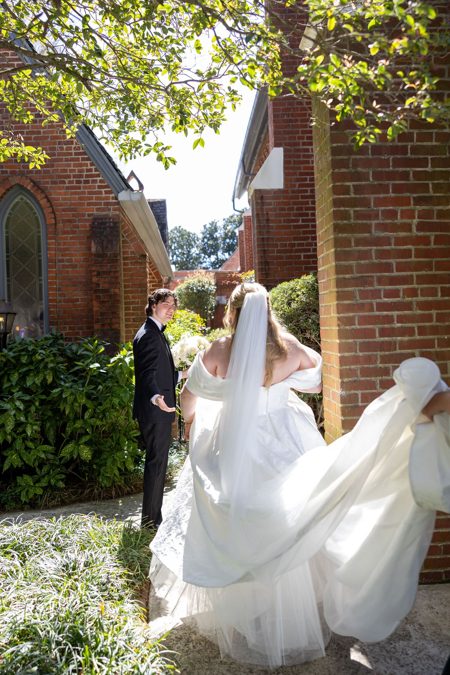 Bride in flowing white gown and veil walks toward groom in black tuxedo outside a brick church on their wedding day.