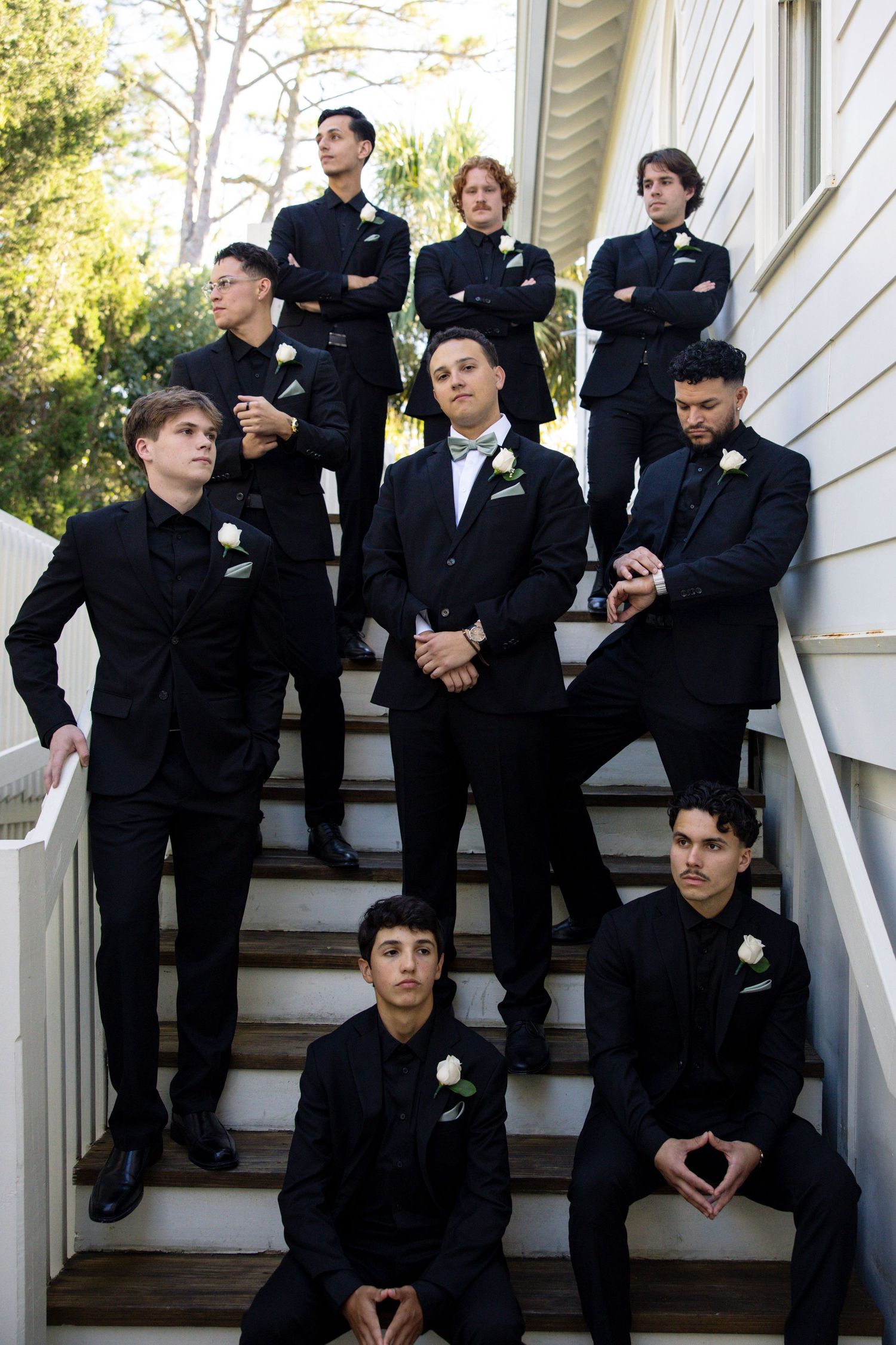 A group wearing matching black formal attire poses together on white wooden stairs outdoors.