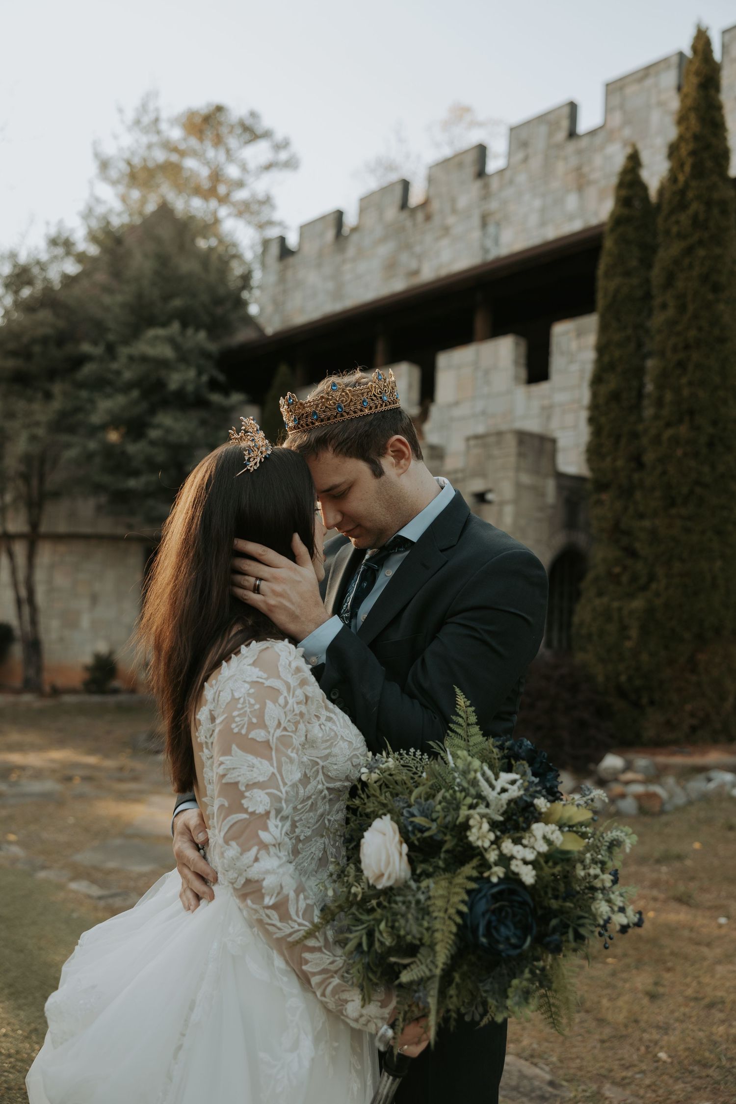 A romantic wedding couple embraces near stone castle walls with cypress trees and floral bouquets at sunset.