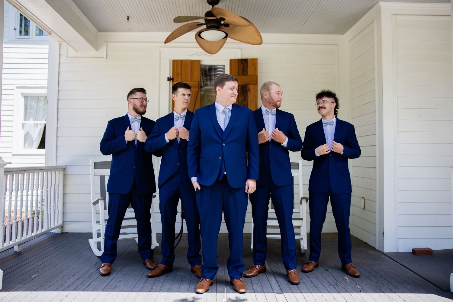 Groomsmen in matching navy suits posing on a white porch with ceiling fan.