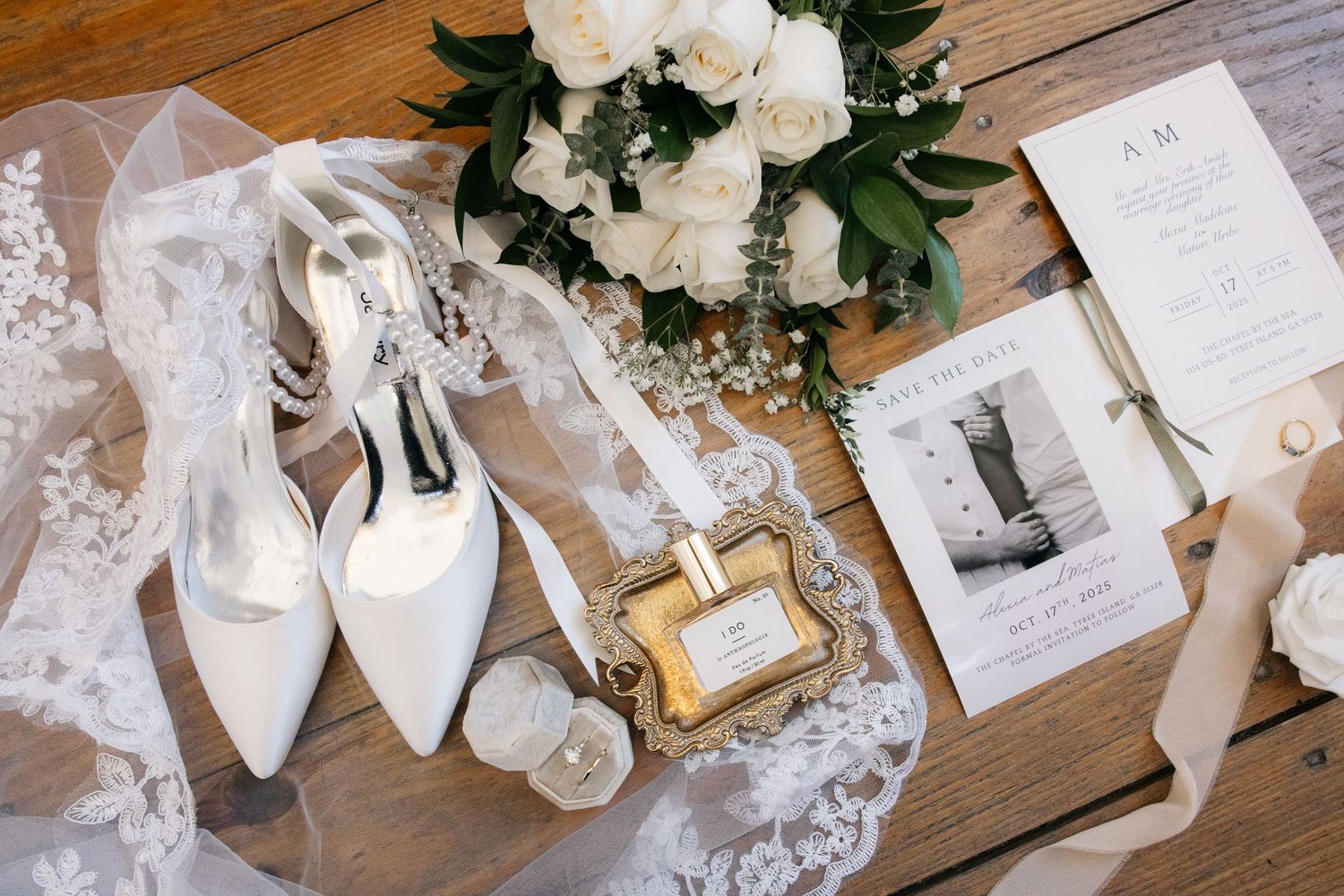 White bridal accessories including lace veil, high heel shoes, invitation cards and floral decor arranged on wooden surface.