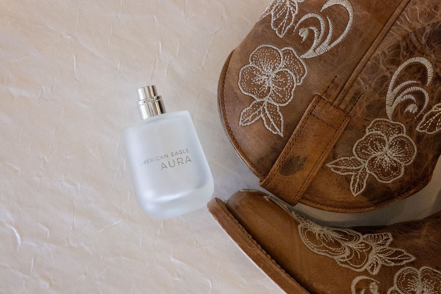 Abercrombie & Fitch Aura perfume bottle beside a brown embroidered leather purse on a white textured surface.