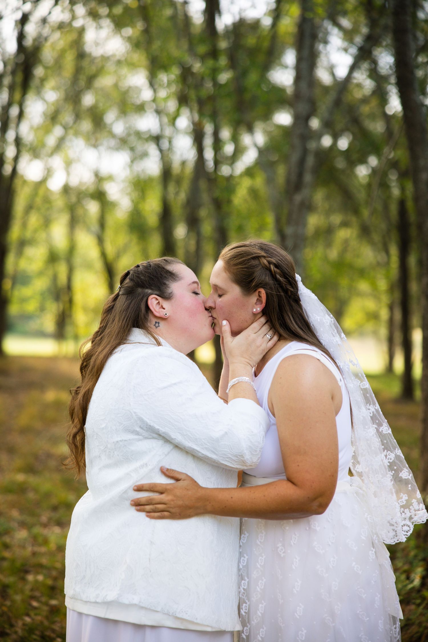 A couple in white wedding attire share a romantic kiss in a wooded outdoor setting.