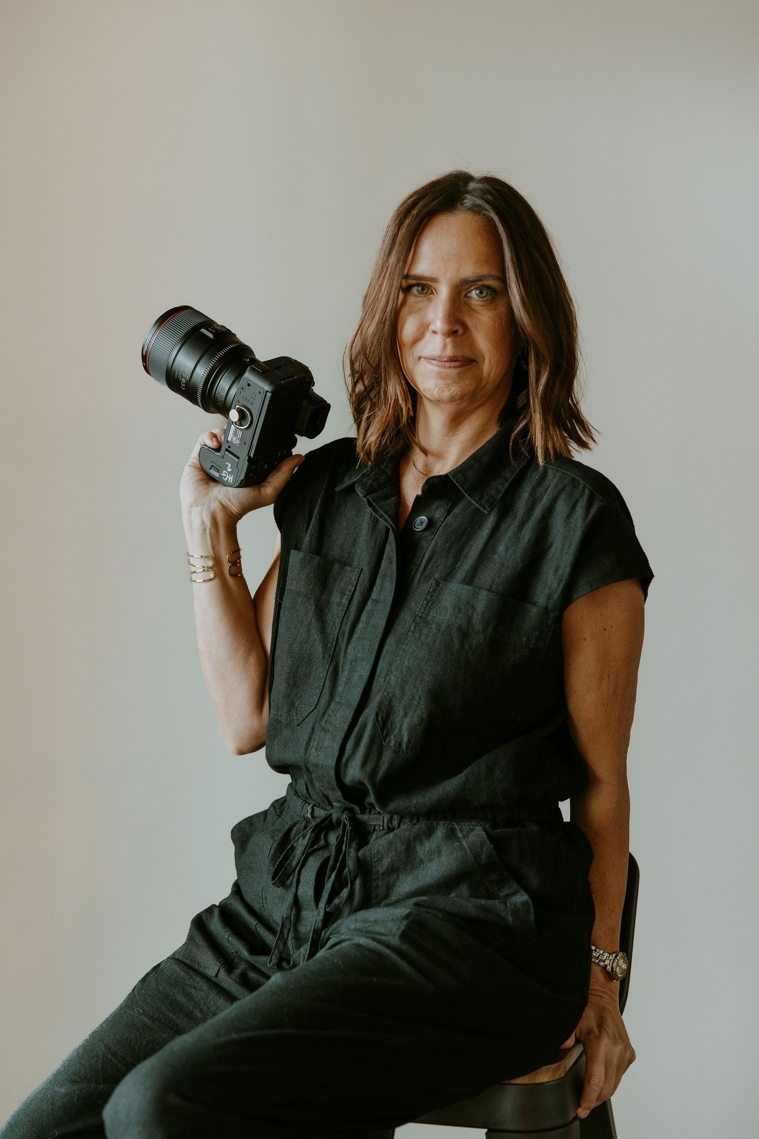 Professional photographer in black outfit holding camera against neutral background.