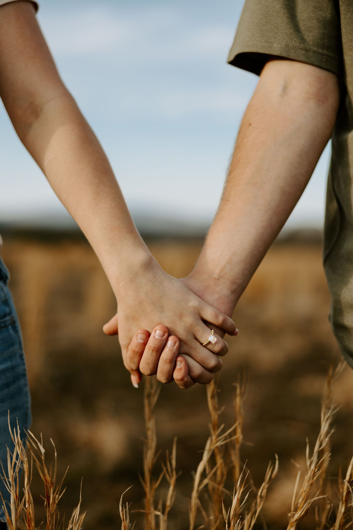 Two people hold hands in a wheat field showing off their wedding rings.
