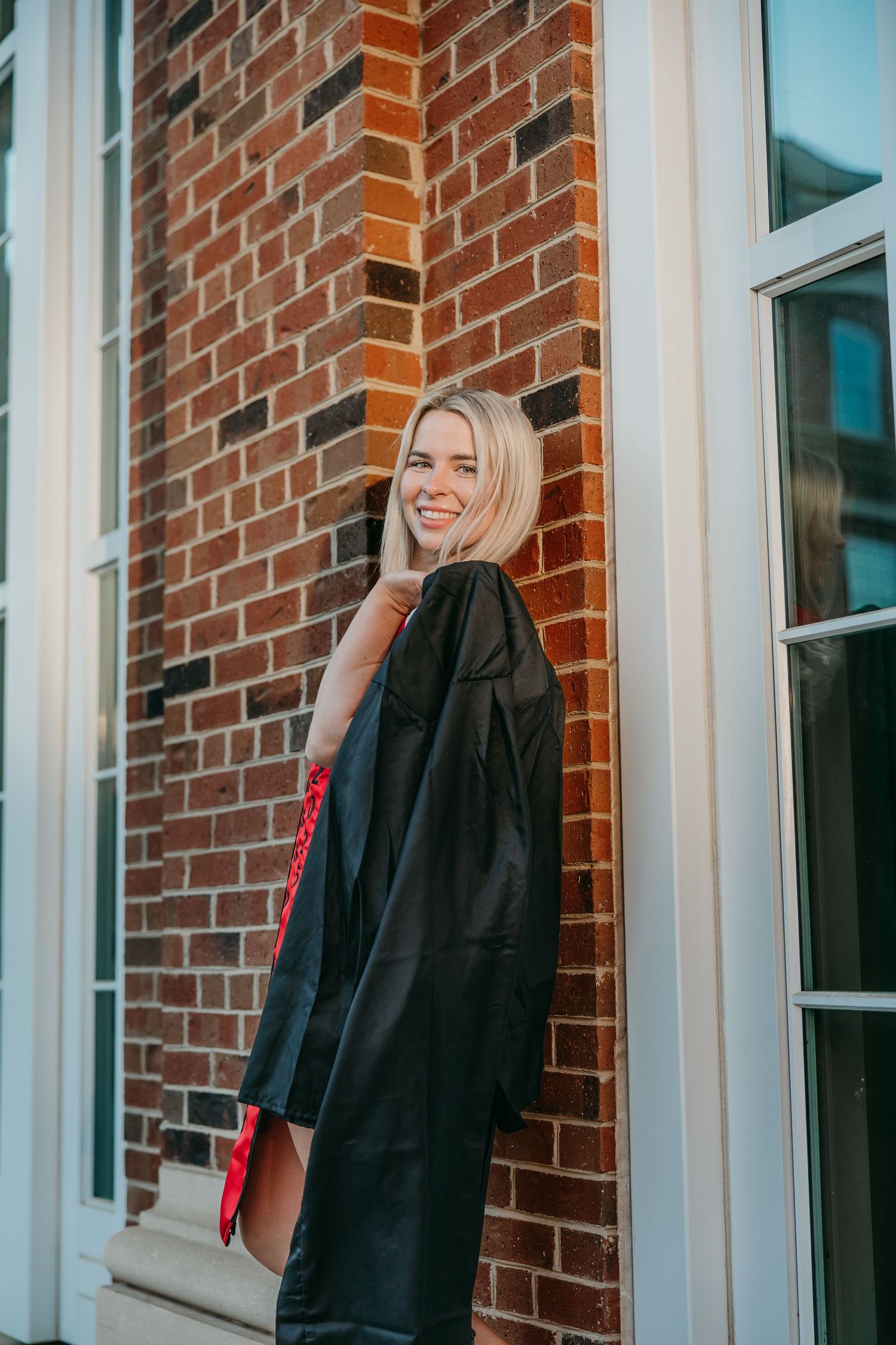 Graduate wearing black gown stands by brick wall smiling.