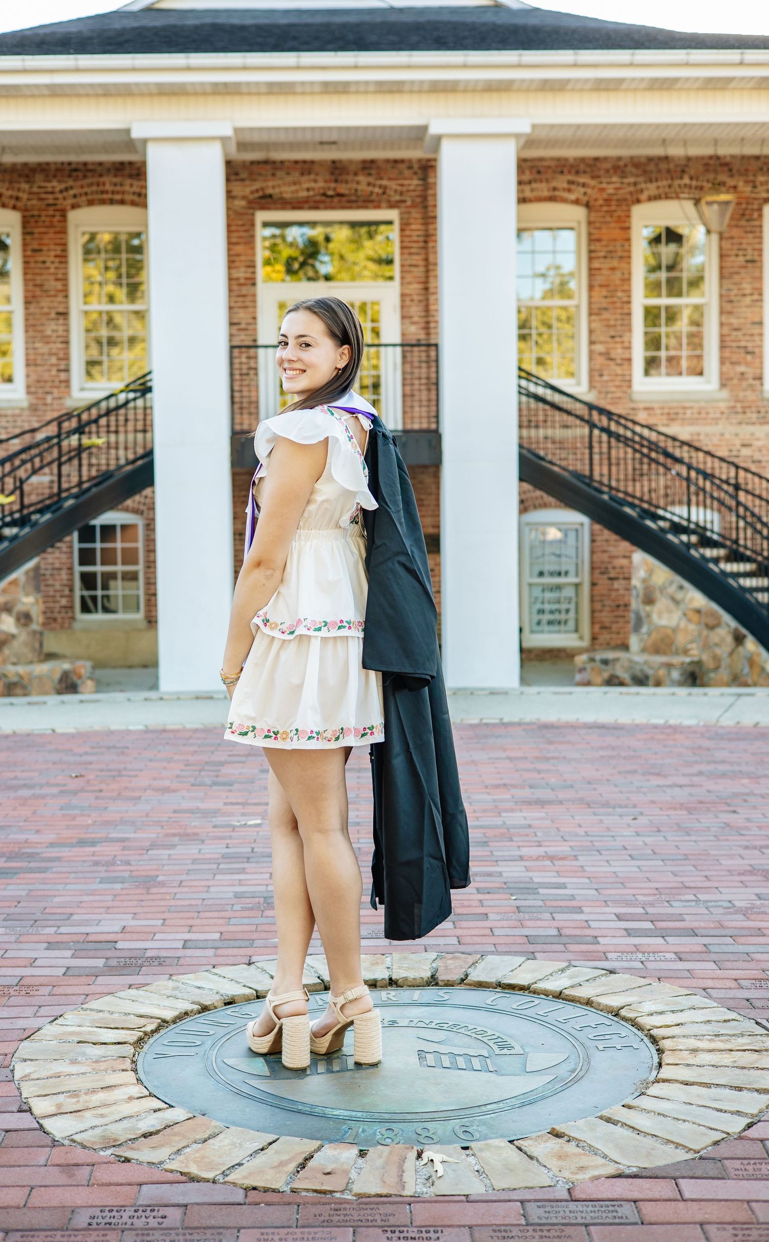 Graduate in white dress poses with cap and gown on decorative circular brick pattern.