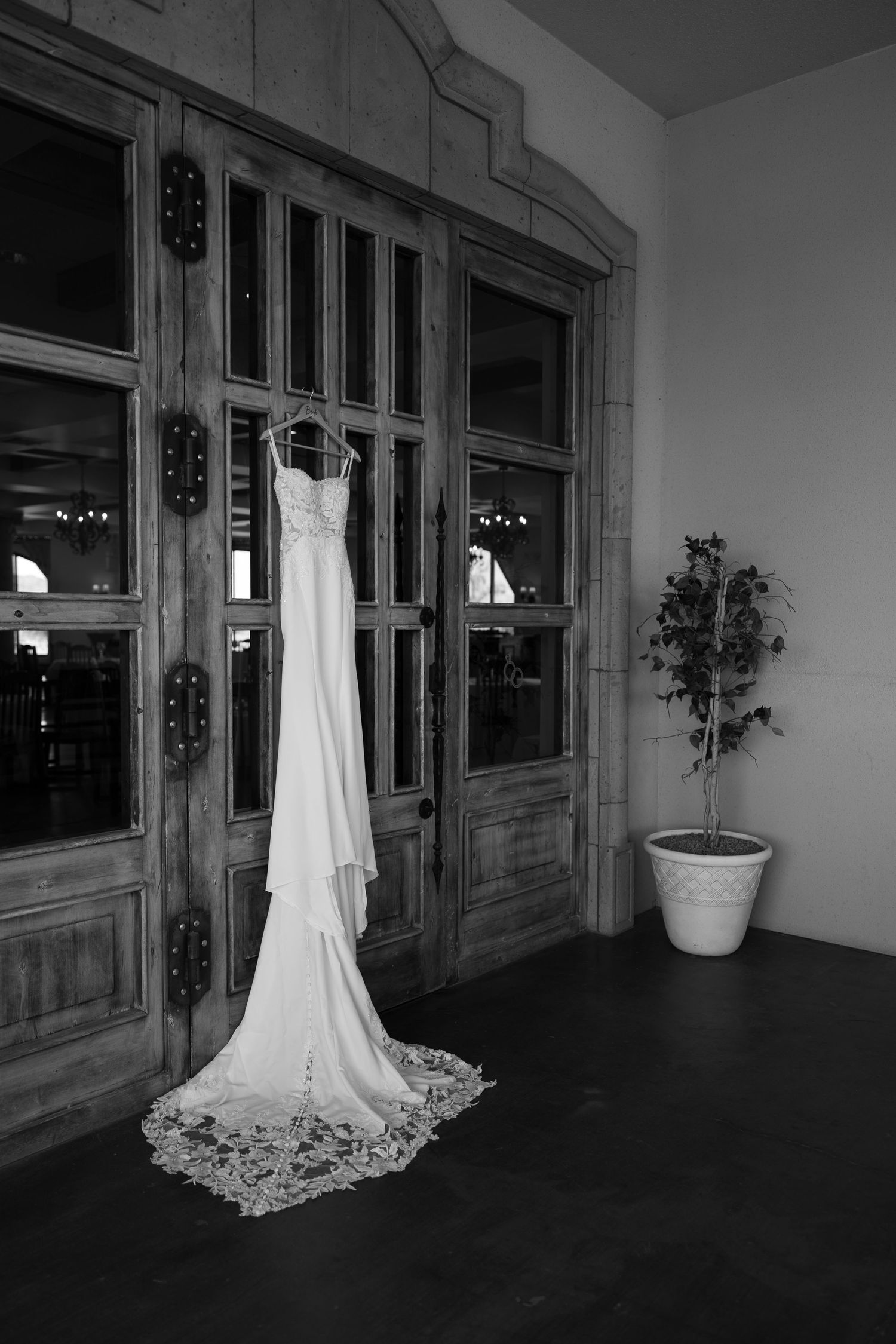 Elegant white wedding dress with lace train hanging on wooden doors in black and white photo.