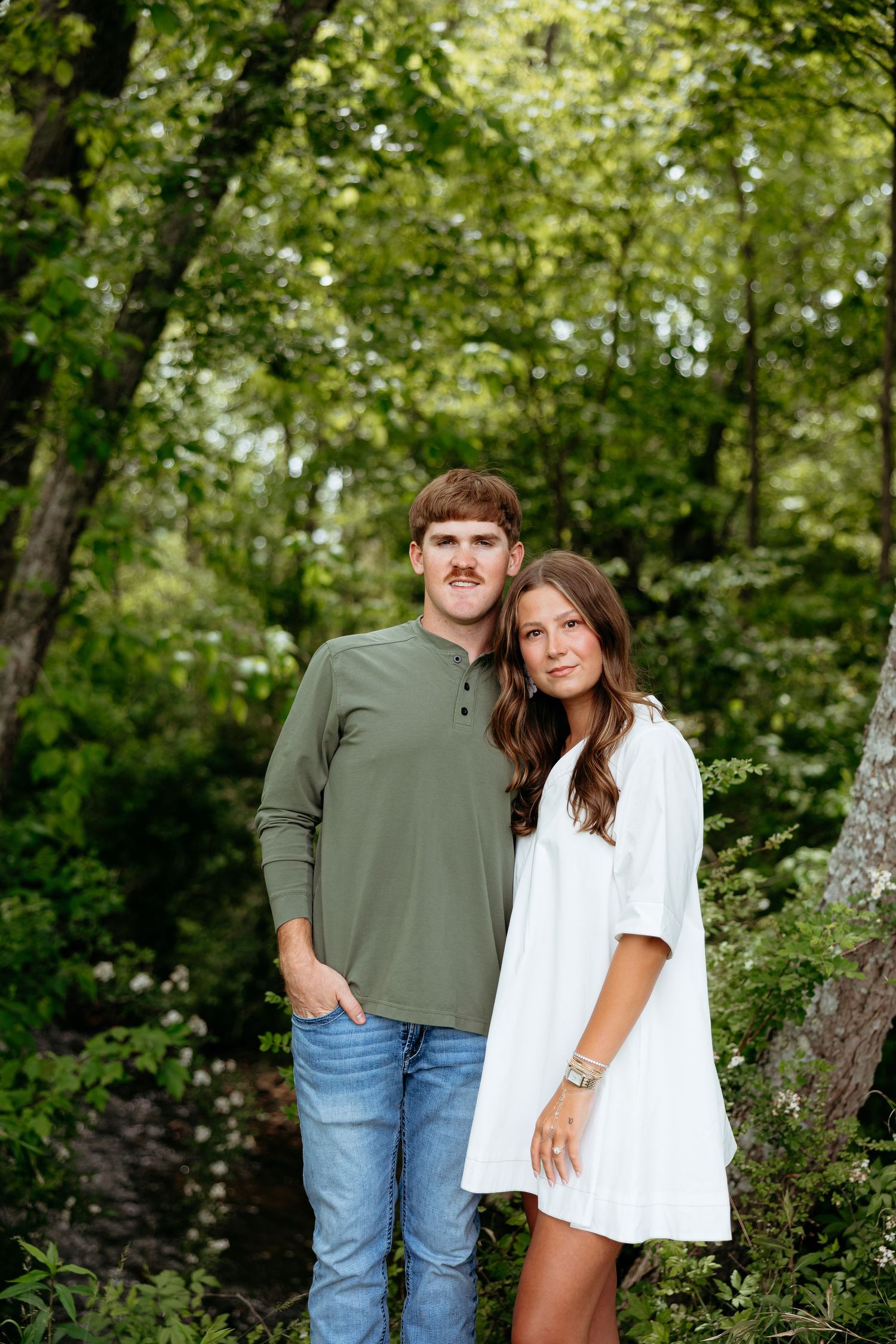 A young couple poses outdoors in a lush green forest, man in olive henley and jeans, woman in white dress.