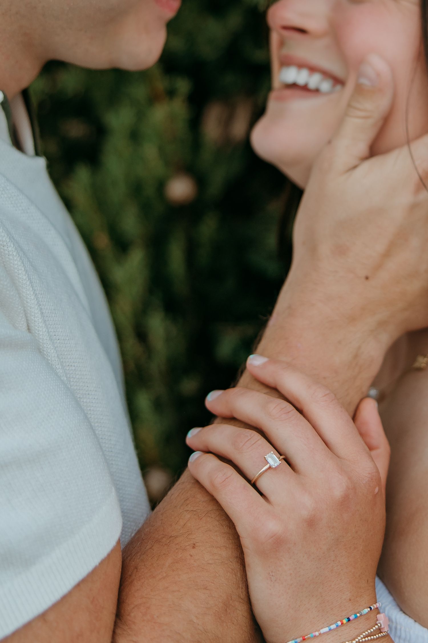 Close-up of hands with engagement ring showing tender moment between couple with natural greenery background.