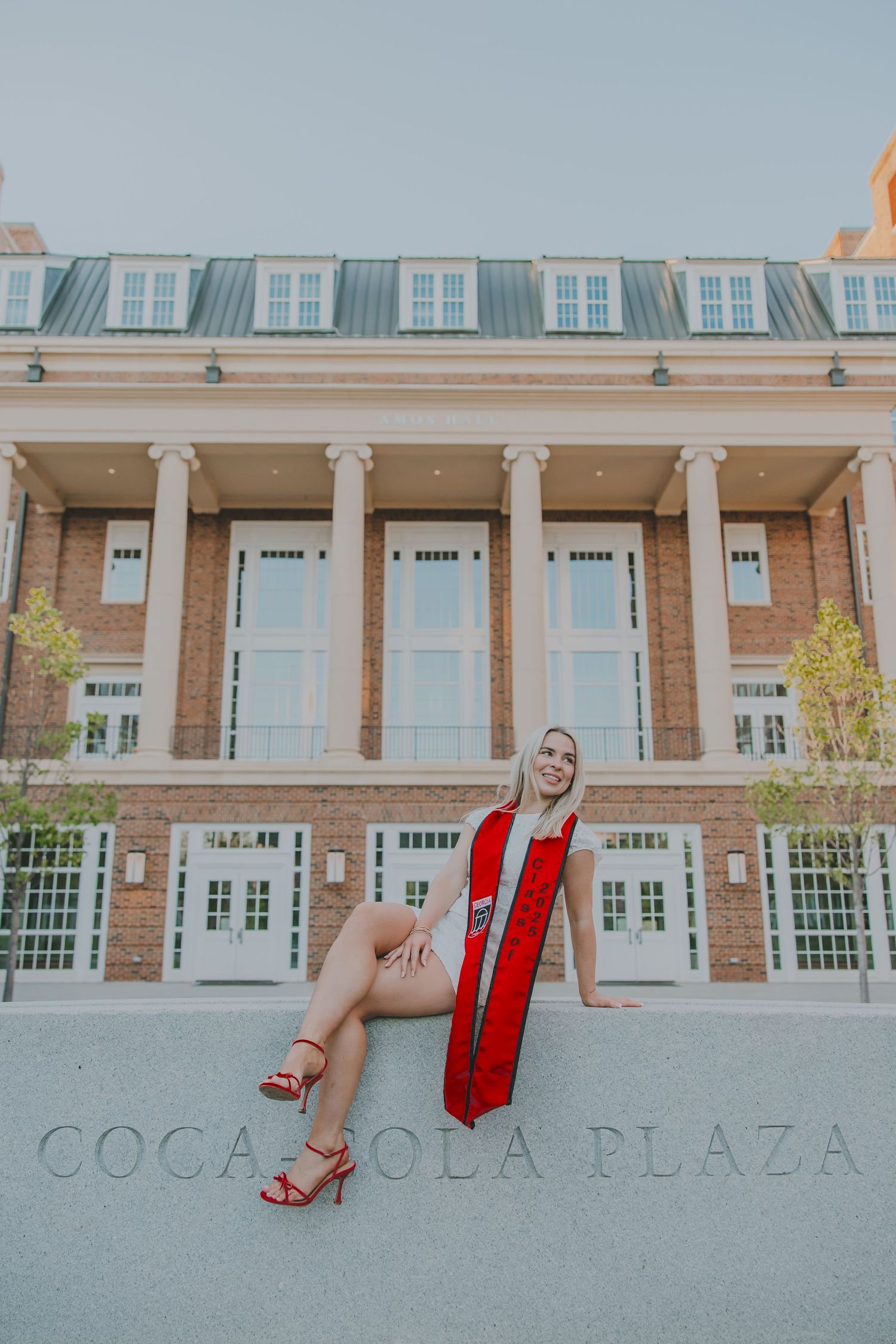 Graduate in red stole poses in front of Coca Cola Plaza building with classical columns.