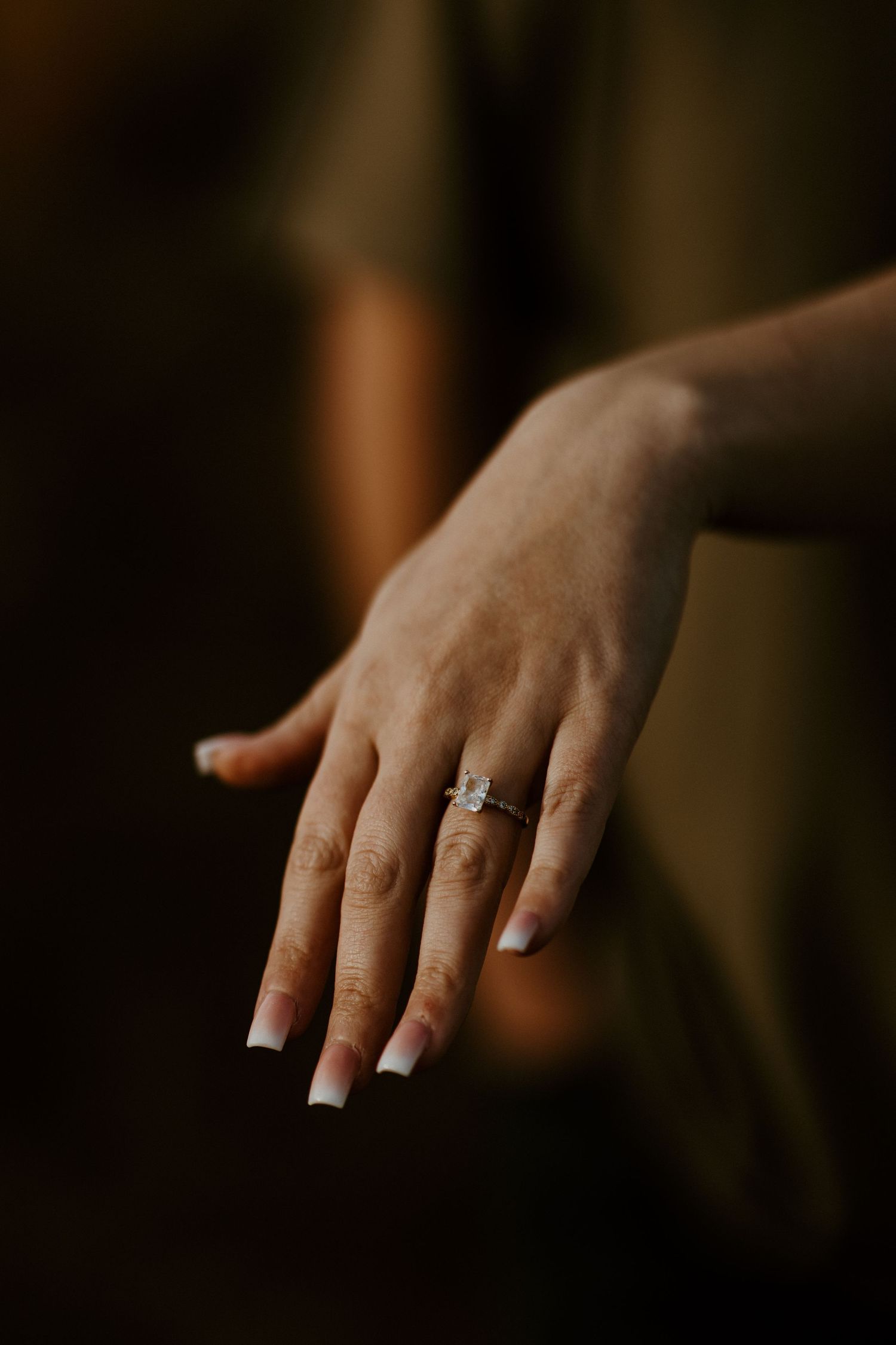 An elegant engagement ring sparkles on a woman's hand with long acrylic nails.