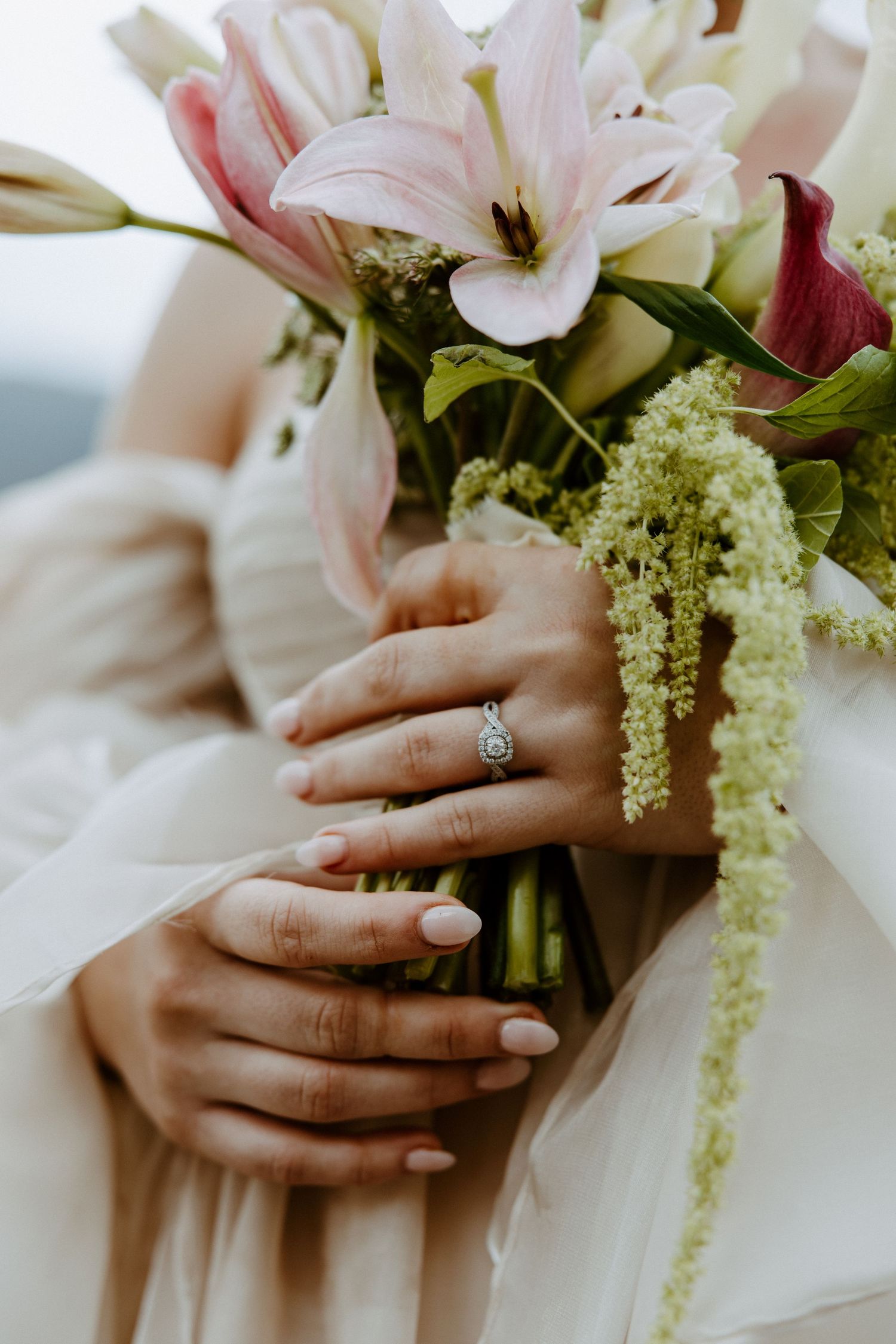 Close-up of hands holding a bridal bouquet with pink lilies and white flowers, showcasing an engagement ring.