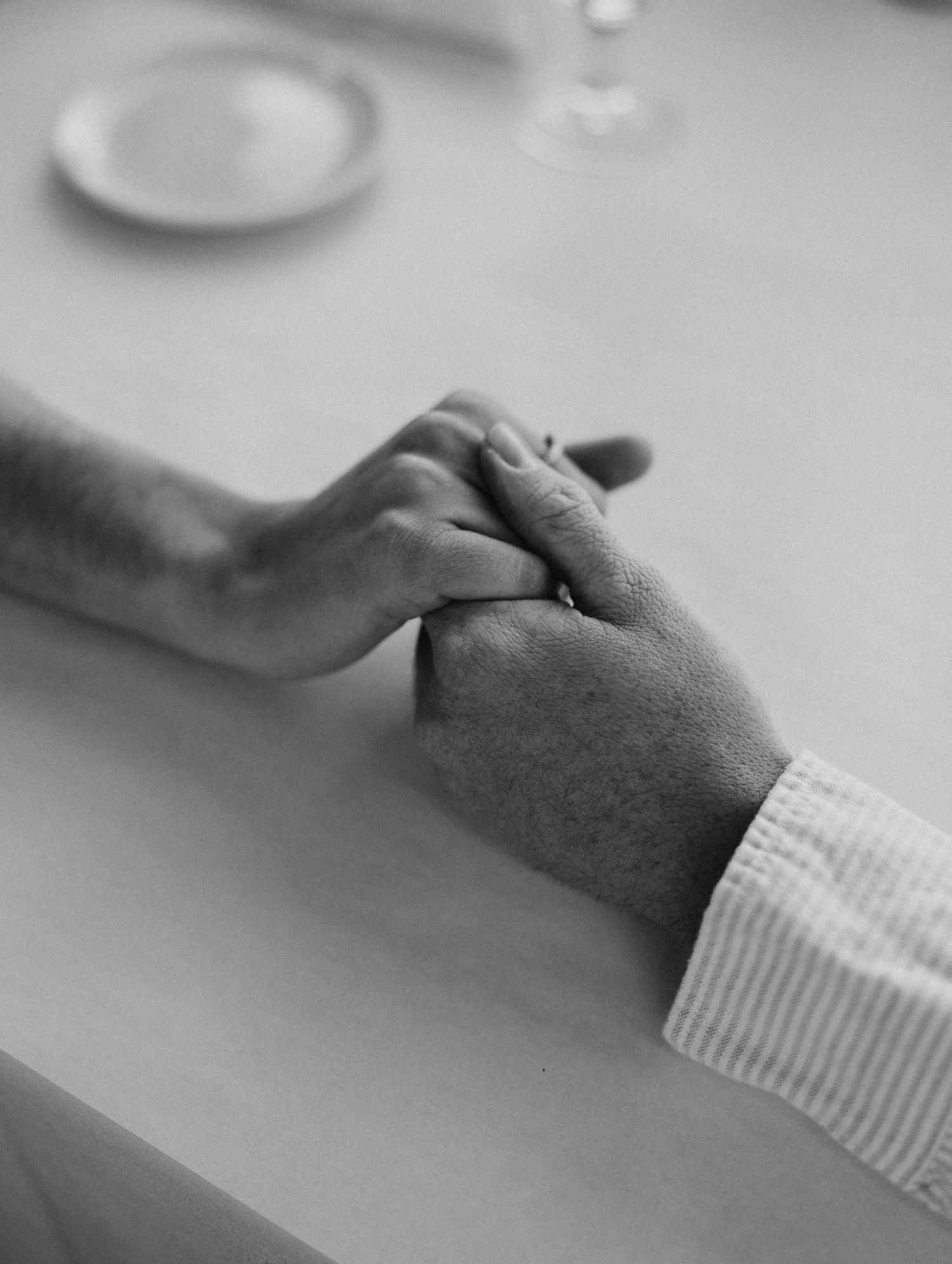 Black and white image of hands tenderly touching across table.