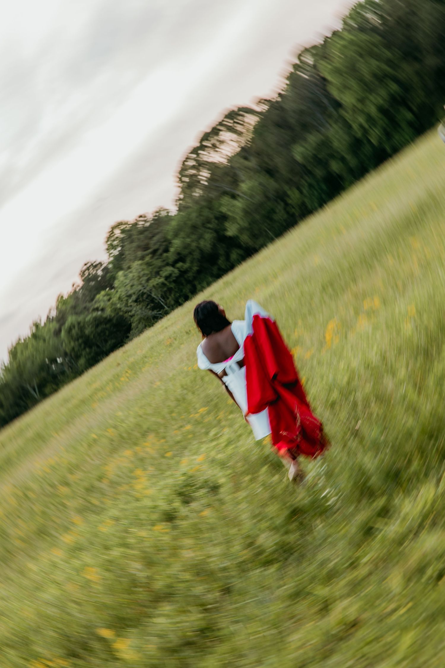 Graduate in red cap and gown walks through a sunny field with trees in the background.