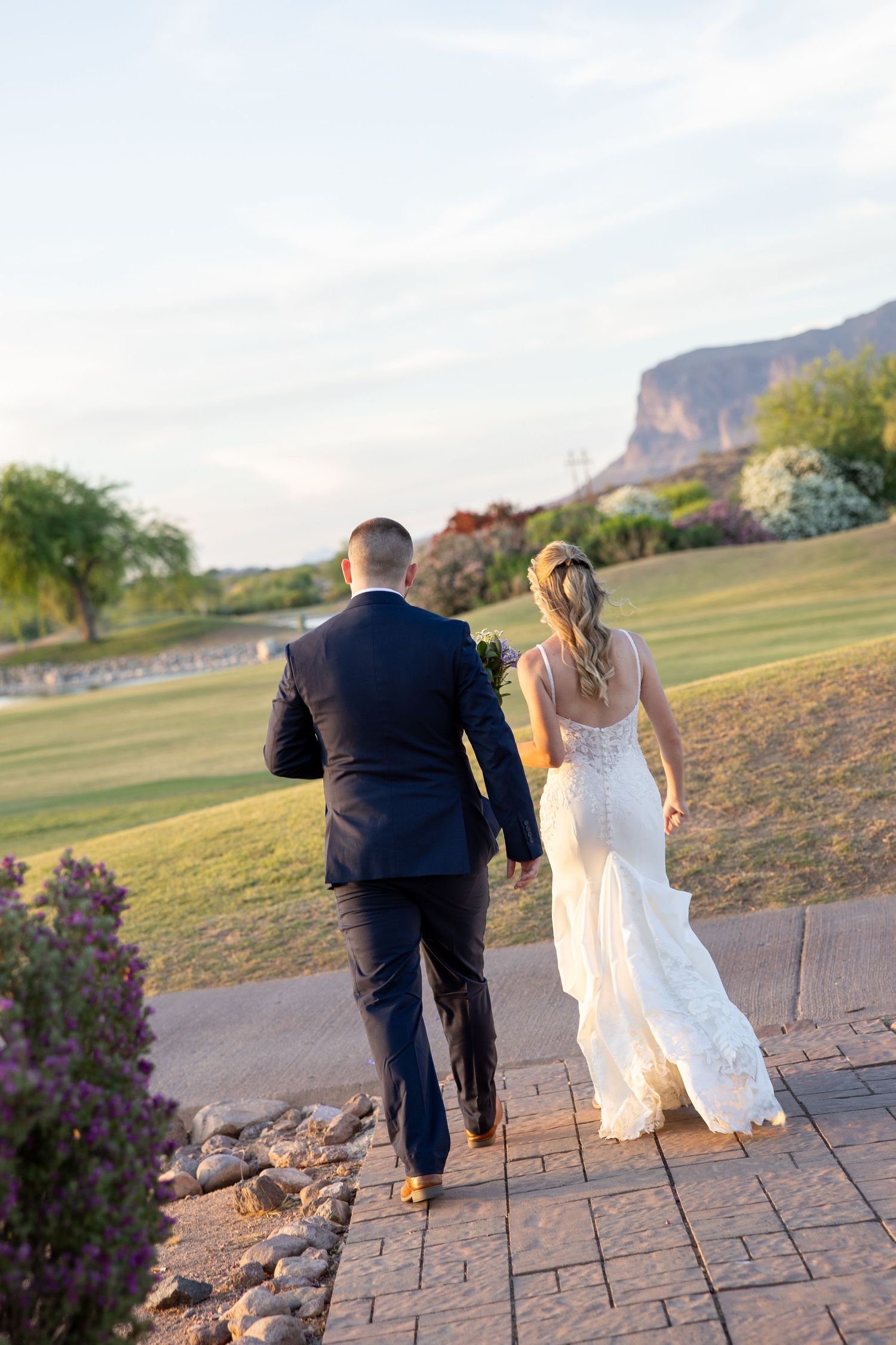 Bride and groom walk hand in hand along stone path toward lush green golf course with mountain views at sunset.