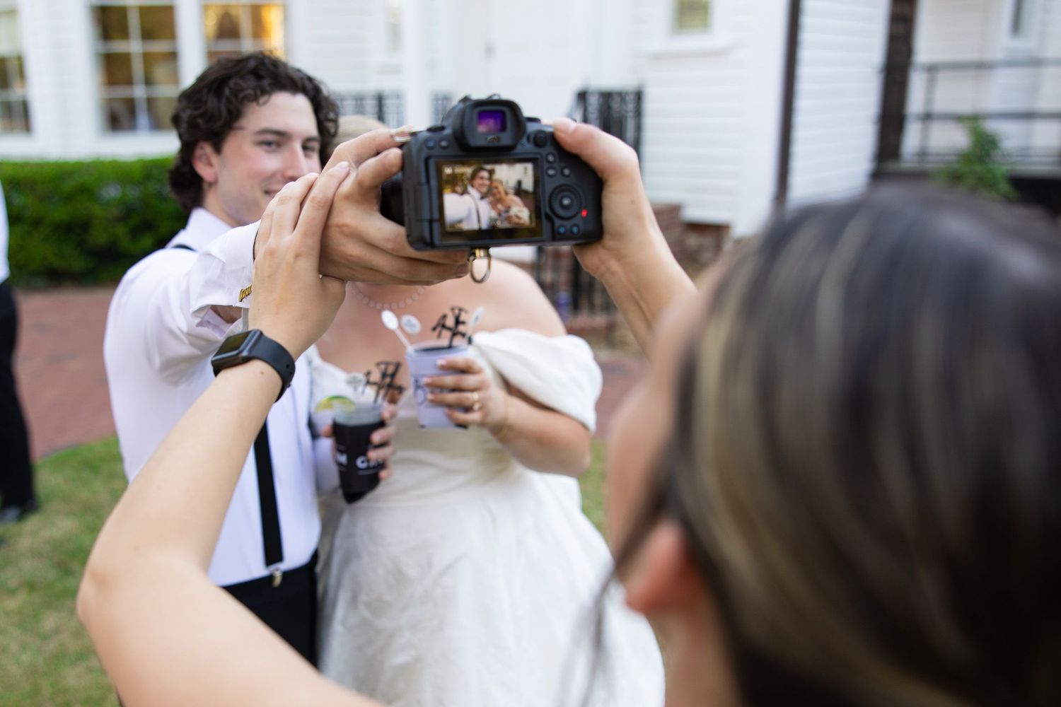 Wedding guests viewing photos on a Canon camera during an outdoor reception with bride and groom in background.