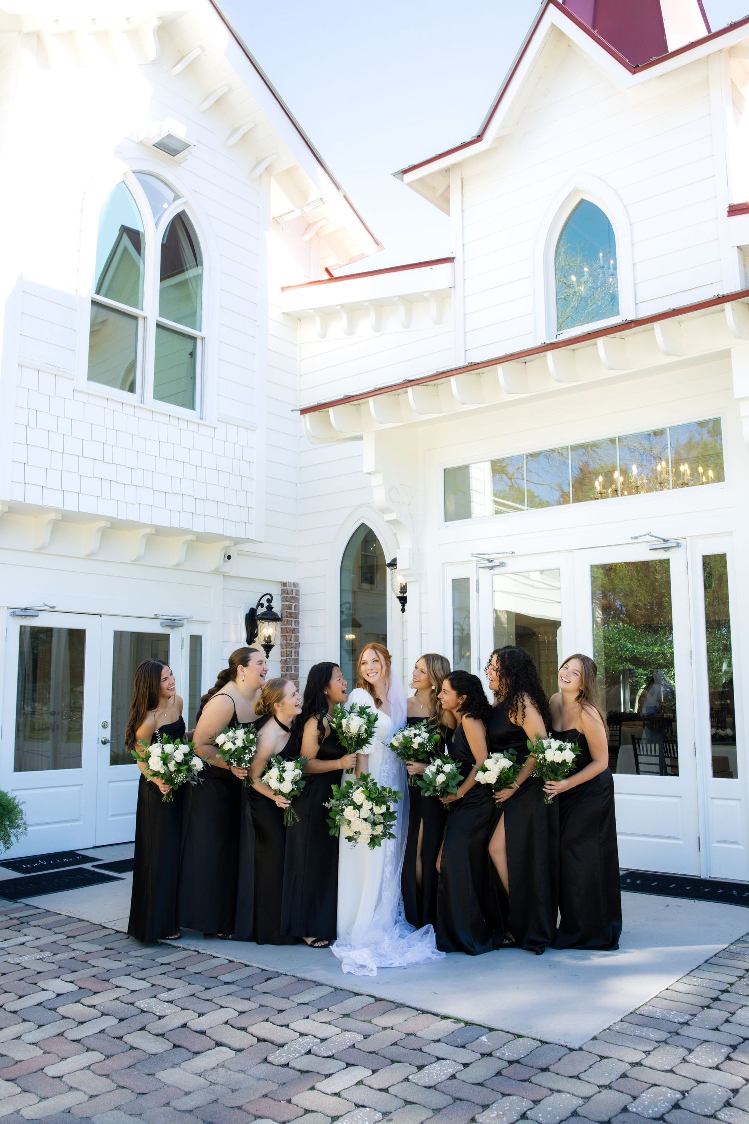 A wedding party in black bridesmaid dresses poses together outside a white chapel with gothic-style arched windows.