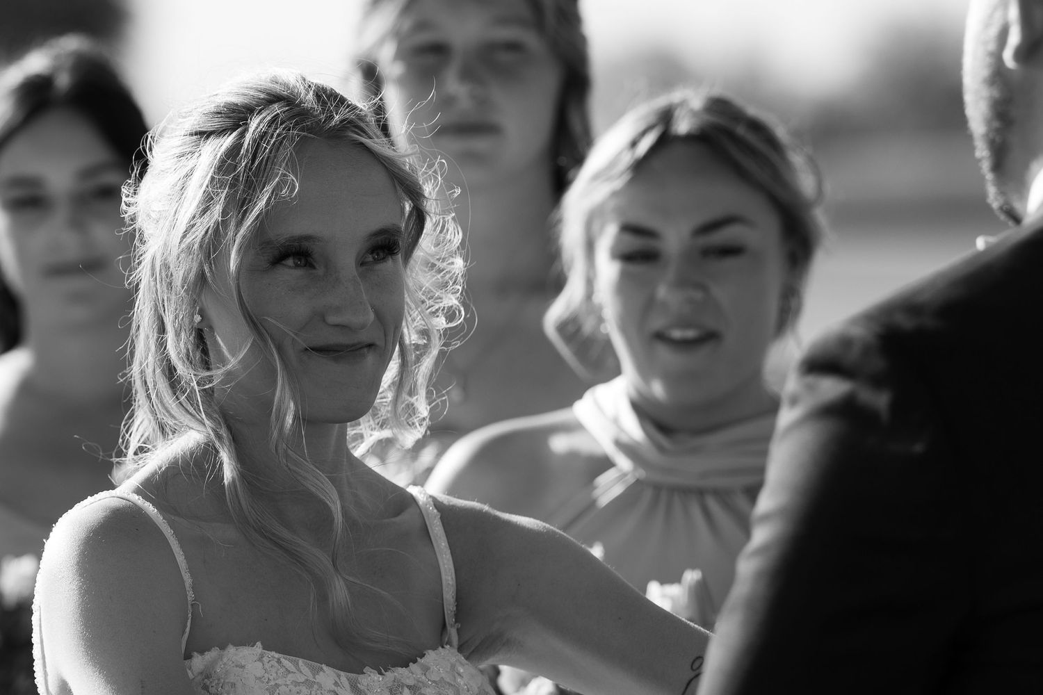 Black and white wedding photo of a smiling bride in lace dress with bridesmaids during outdoor ceremony.