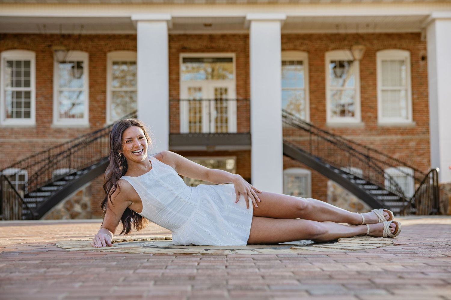 Graduate in white dress reclining on brick courtyard for portrait.