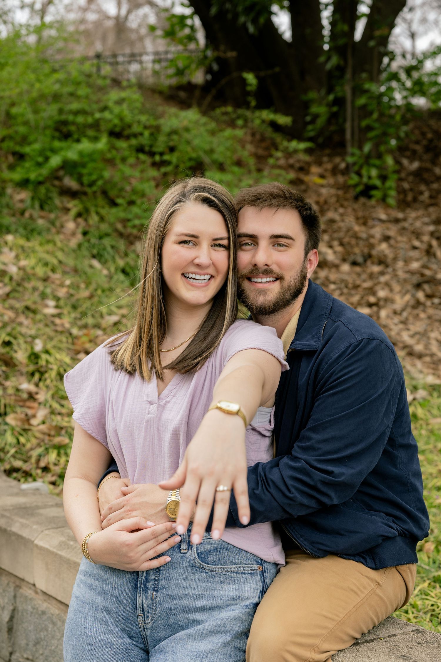 A newly engaged couple poses together outdoors showing off an engagement ring.
