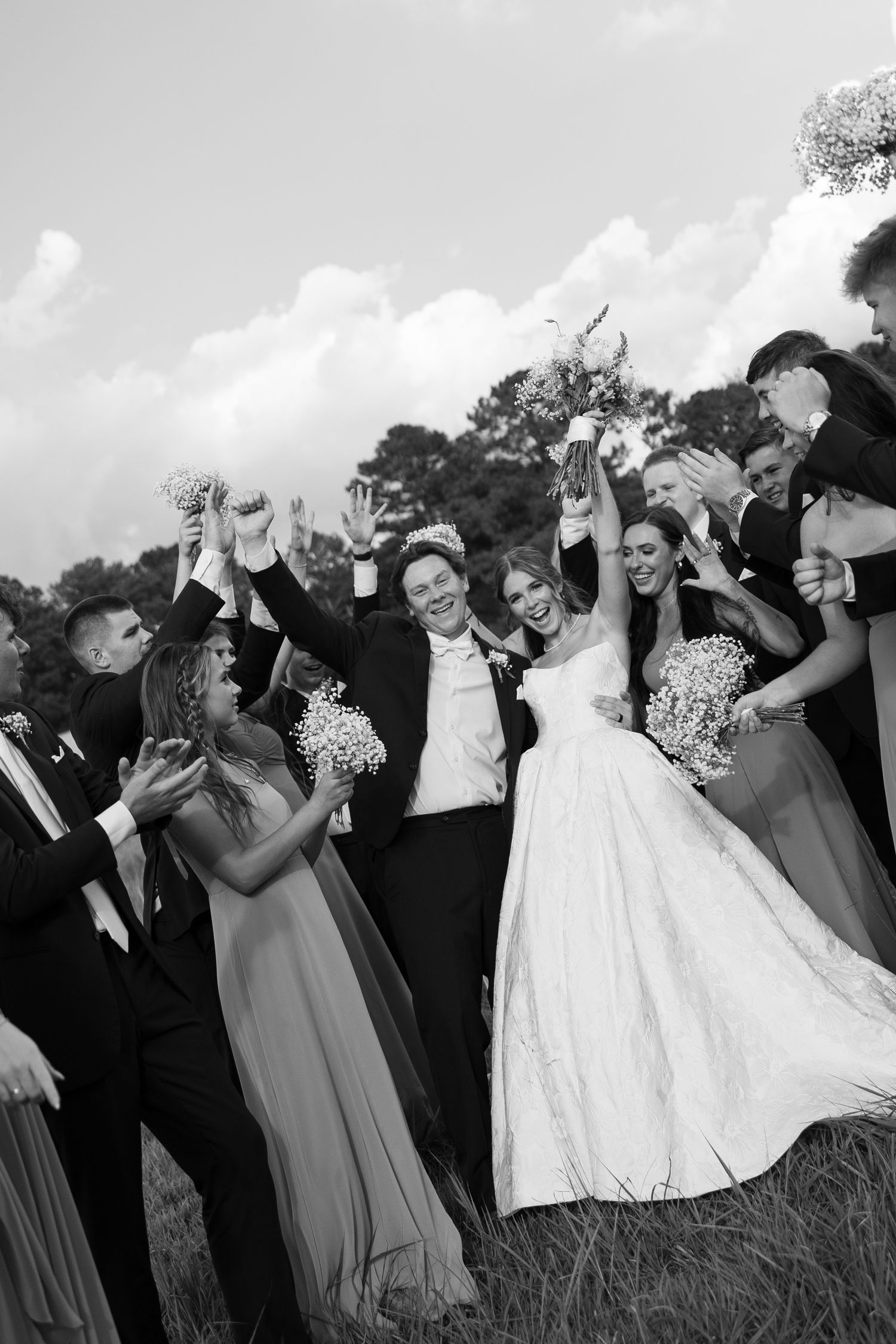 Wedding party celebrates with arms raised in a joyful dance line outdoors in black and white photography.