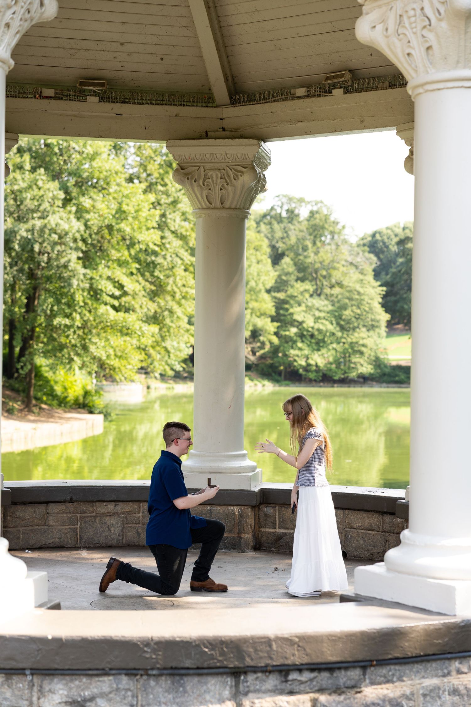 Man proposing on one knee to woman in white dress under pavilion columns by serene lake in park.