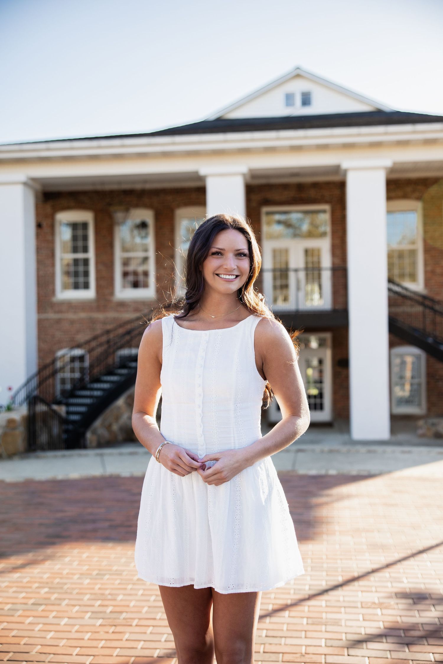 Graduate in white summer dress smiling in front of campus building with columns.