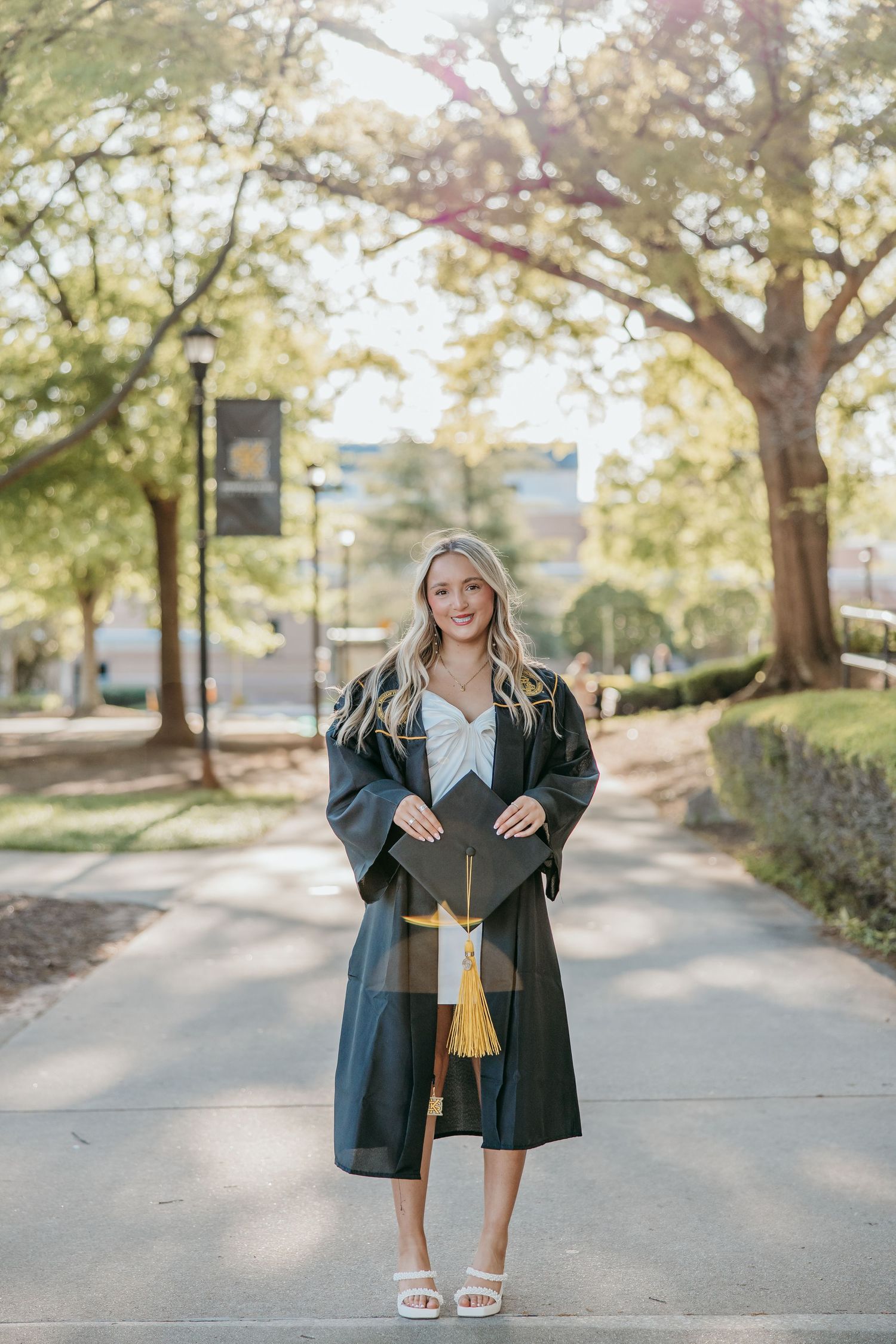 Graduate stands on campus path wearing traditional dark graduation gown and cap.