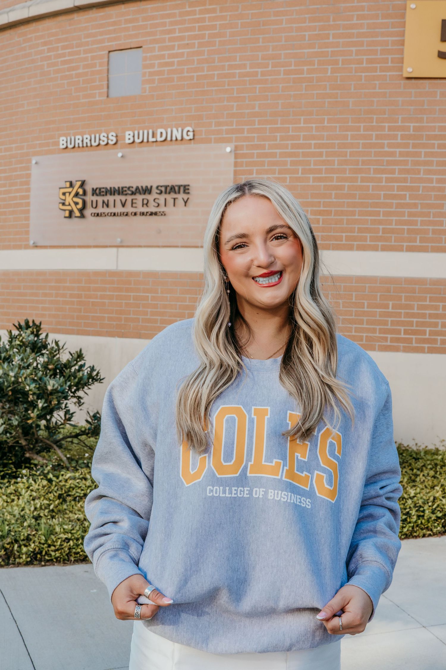 College student poses in light blue COLES sweatshirt outside Burgess Building at Kennesaw State University.