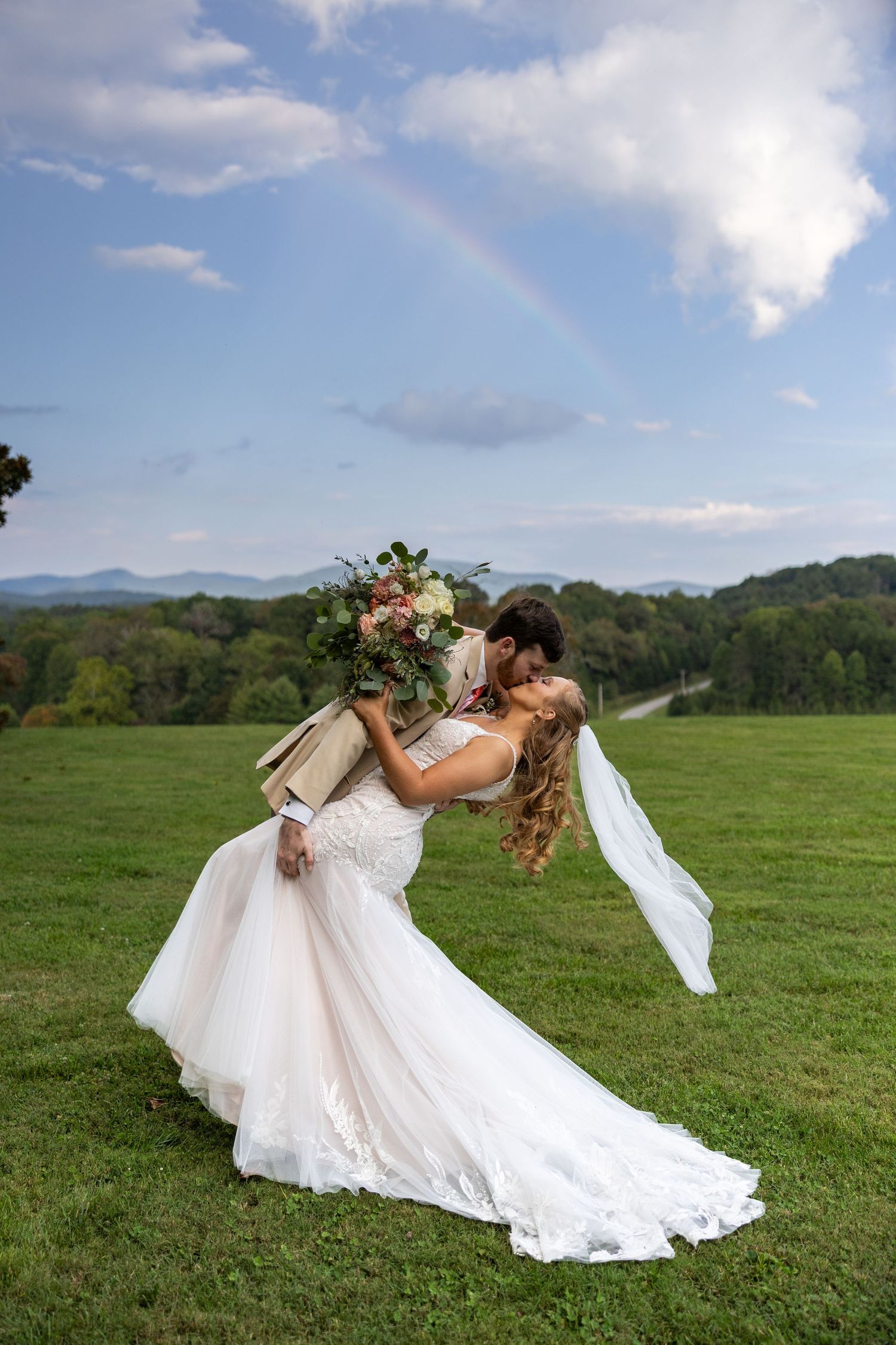 A romantic wedding couple shares a dip kiss in a scenic mountain field with a flowing white dress and blue sky.