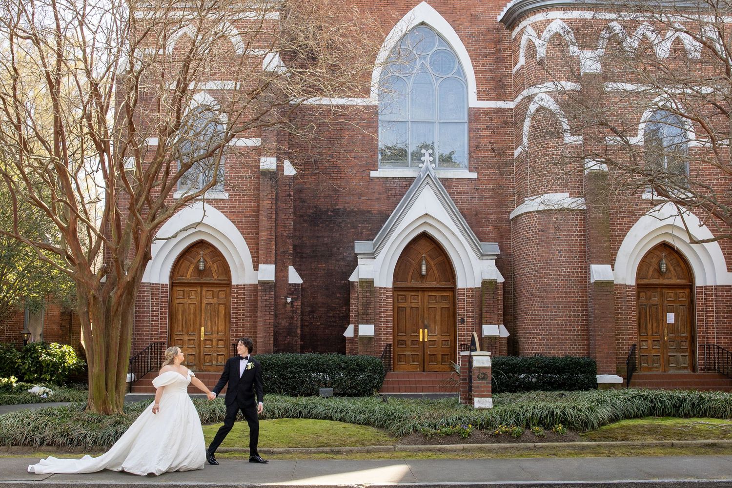 A wedding couple walks together outside a gothic-style brick church with arched windows and doorways.