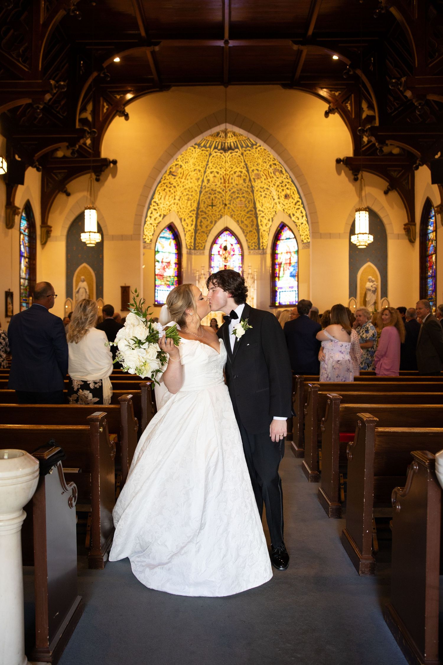 Bride and groom share a kiss in the aisle of a beautiful church with stained glass windows after their wedding ceremony.