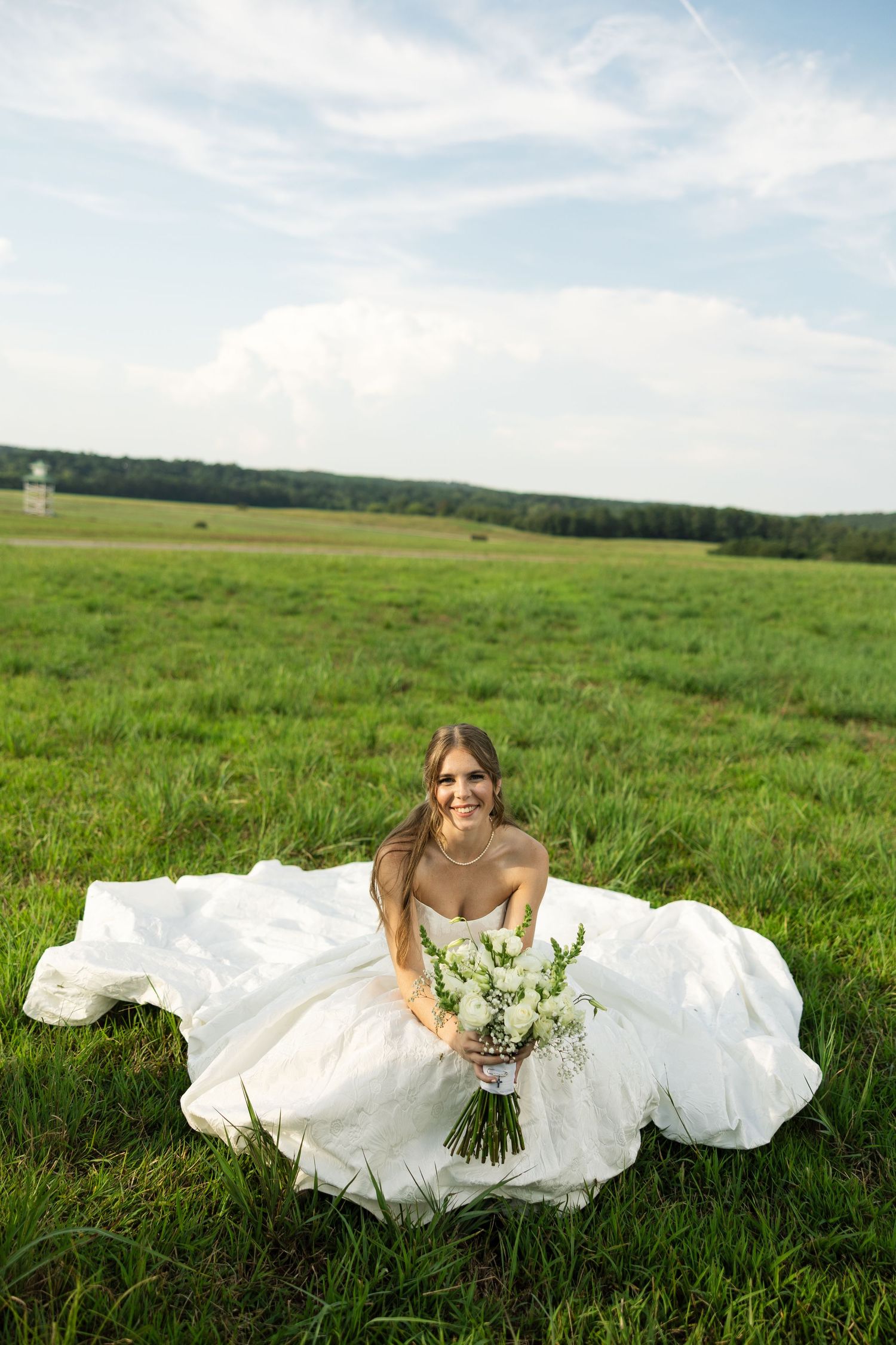 A bride in a flowing white wedding dress lies gracefully in a lush green meadow under blue summer skies.