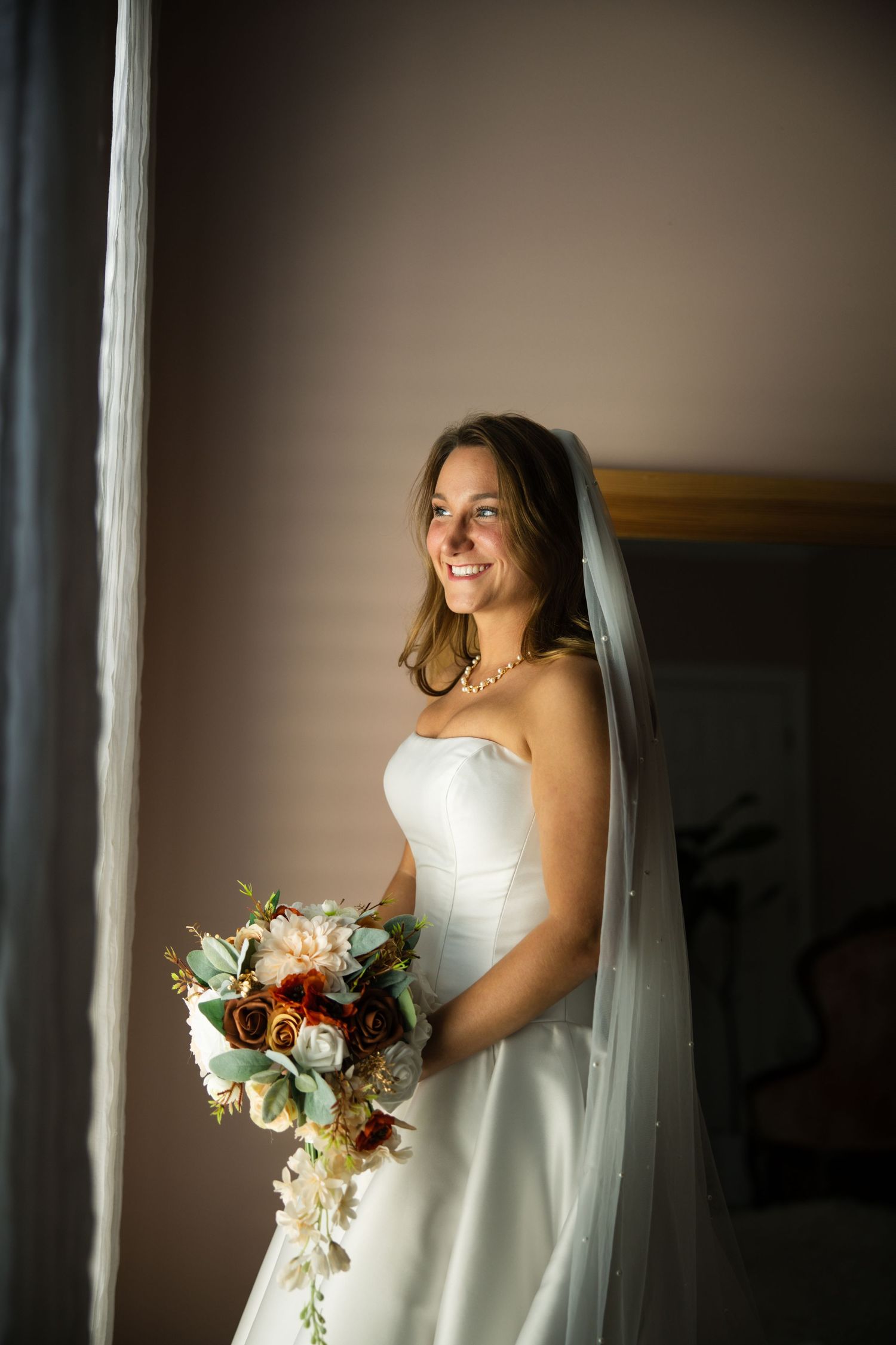 A bride in a white wedding dress stands near a window holding a beautiful bouquet of mixed flowers in soft lighting.