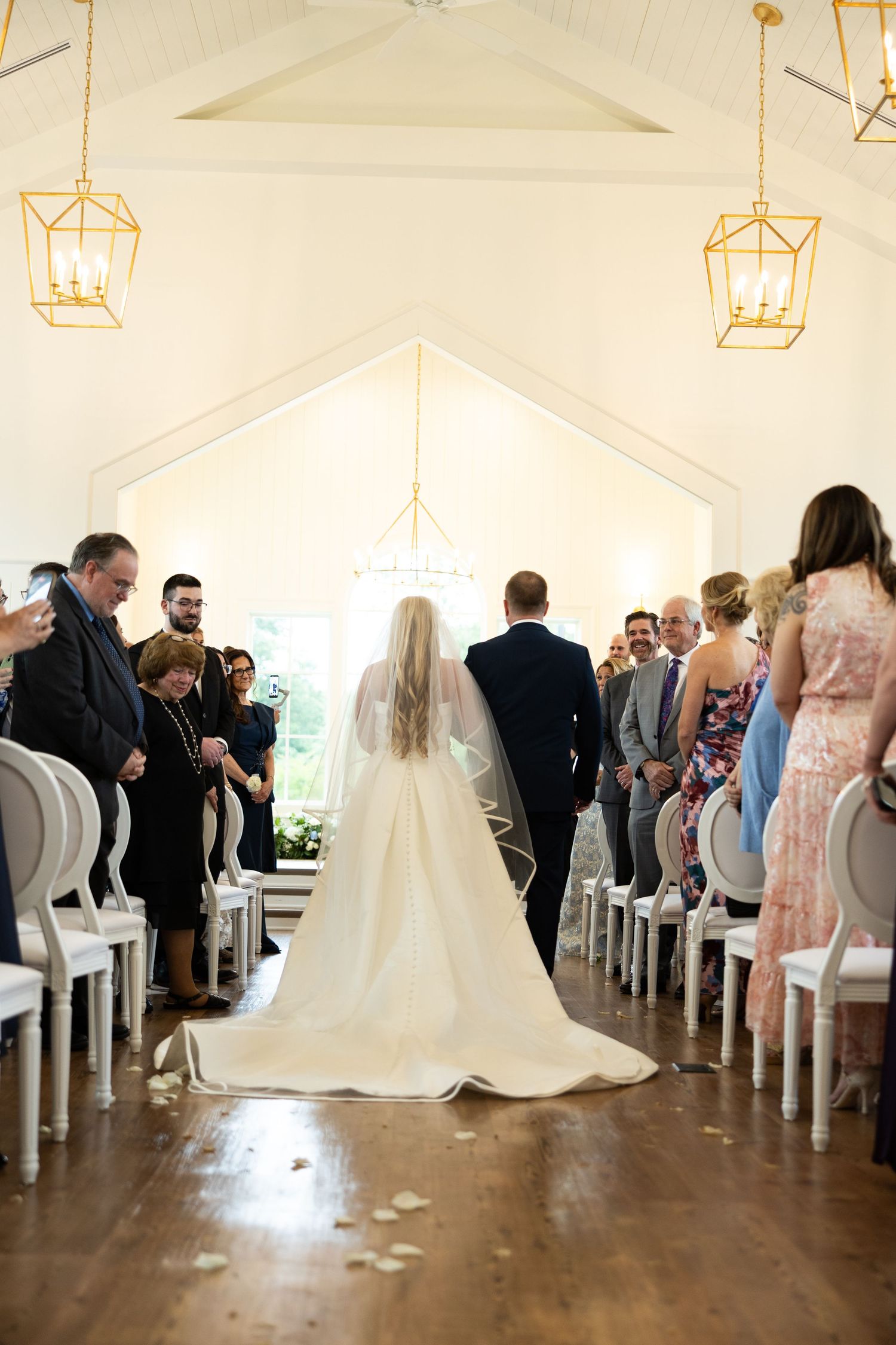 Bride walking down the aisle in a white gown and veil in an elegant chapel with gold lanterns.